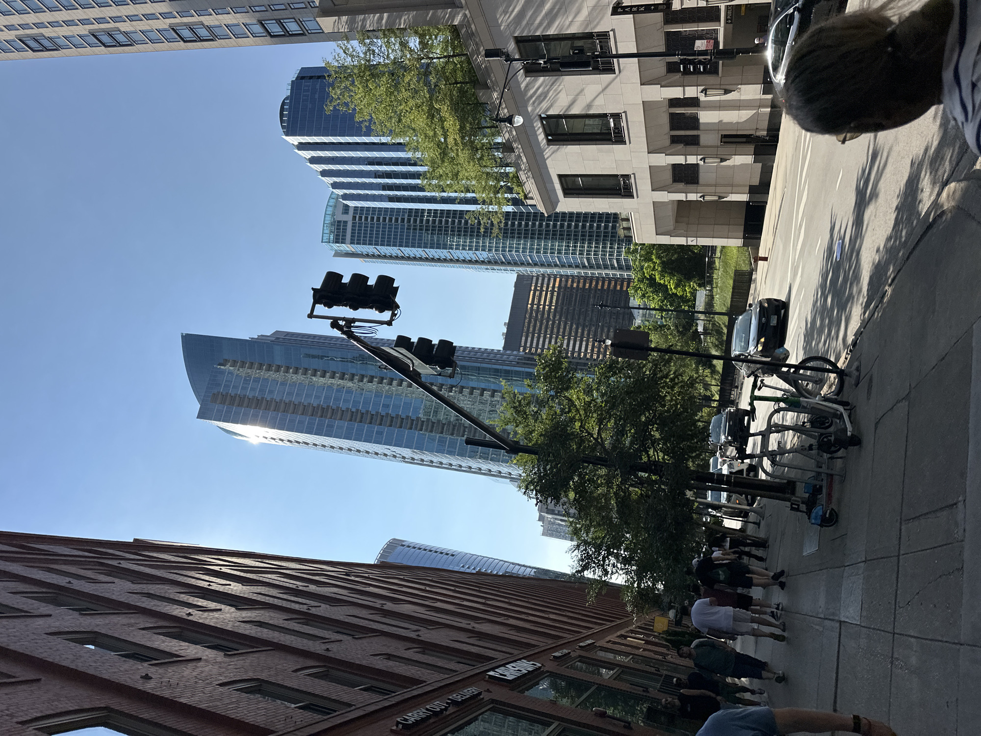 This image depicts a street scene in Chicago, USA, on a clear, sunny day. The perspective is from a sidewalk looking down a street lined with modern skyscrapers. On the left, a red brick building with numerous windows occupies the foreground, its facade angled towards the sky. To the right, several tall, glass-fronted buildings rise, one notably curved. A traffic light with three dark signals hangs above the street.
On the sidewalk, several individuals are walking, some in casual attire, and two bicycles are parked near a rack. In the street, a black sedan is visible. The environment suggests an urban setting with significant architectural development.