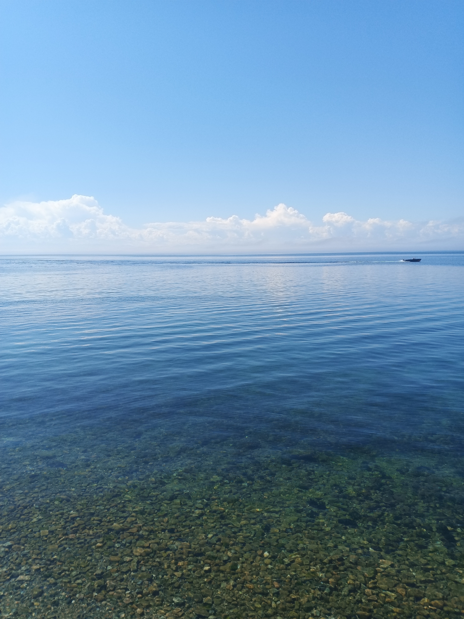 This image captures a serene view of Lake Baikal in Listvyanka, Russia. The upper portion of the frame is dominated by a clear, bright blue sky with scattered white, puffy clouds forming a distinct band across the horizon. Below the sky, the vast expanse of Lake Baikal stretches out, its surface reflecting the blue of the sky and exhibiting subtle ripples from a gentle breeze. The water transitions from a deep blue in the distance to a lighter, more transparent hue closer to the shore. In the foreground, the lakebed is visible, covered in smooth, rounded pebbles and rocks of various sizes and colors, including browns, greens, and grays. A small, dark-colored boat is visible in the distance, moving from right to left, leaving a faint wake on the water. The overall scene conveys a sense of tranquility and natural beauty on a sunny day.
