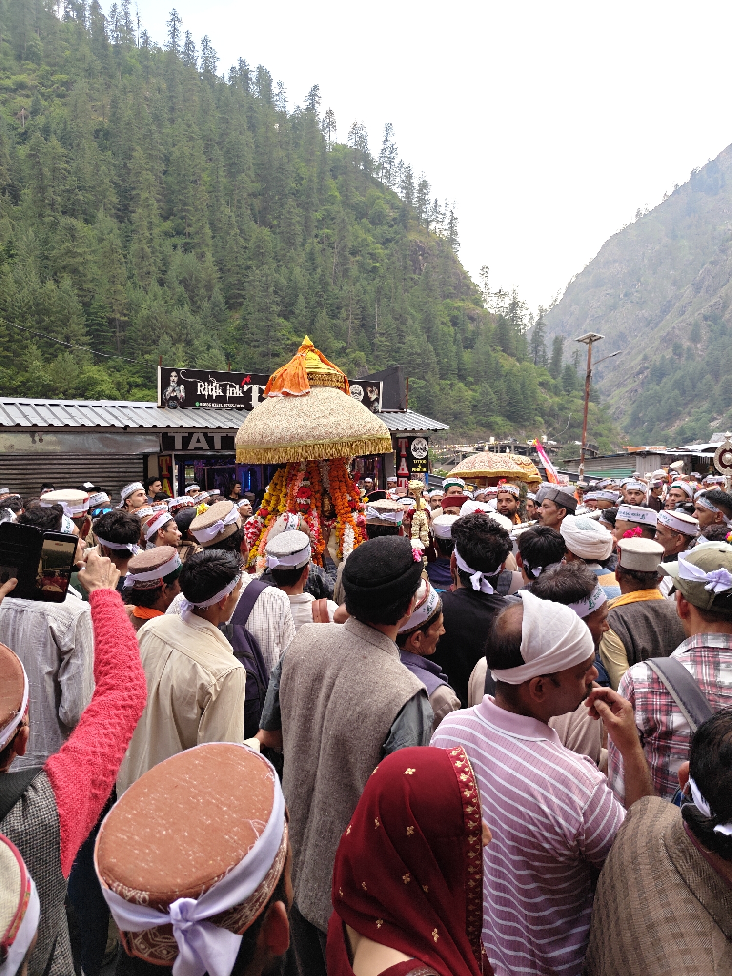 This image captures a vibrant and crowded scene in Manikaran, India, set within a narrow valley flanked by steep, densely forested mountains. The central focus is a richly decorated palanquin or chariot, adorned with colorful fabrics (gold, orange, and white/cream) and garlands of flowers, likely carrying a deity. It features a distinctive, ornate umbrella-like top.
A large crowd of people, predominantly men, is gathered around this central structure, participating in what appears to be a religious procession or festival. Many individuals in the crowd wear traditional Himachali caps, often round with patterned bands or simple white cloth wraps, signifying the local culture. Some people are seen holding up mobile phones, capturing photos or videos of the event.
In the background, behind the procession, are several low-rise buildings with corrugated metal roofs, housing shops or stalls. A prominent black signboard reads "Ritik Ink" with the services "TATTOO & PIERCING" and phone numbers "93686 83331, 97365-11035" visible. Part of another sign says "TAT". A street light with solar panels is also visible on the right side of the frame.
The surrounding environment is characterized by towering mountains covered in thick coniferous forests, establishing a distinctly Himalayan setting. The sky is overcast, suggesting a cloudy day, but there is ample daylight, indicating it is likely morning or early afternoon. The atmosphere appears to be one of communal gathering and engagement with the event.