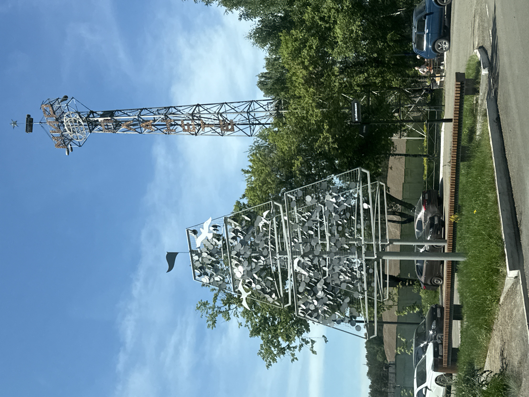 The image shows two metal sculptures in Chaikovsky, Russia. One is a tall, open framework tower topped with a star. The other is a multi-tiered, geometric structure filled with round, reflective discs and stylized bird figures. Both are located near a street, with cars parked alongside the road, and trees in the background. The sky is blue with some clouds. A street sign reading "Ул. Космонавтов" is visible. Two people are visible between the trees and a blue car.