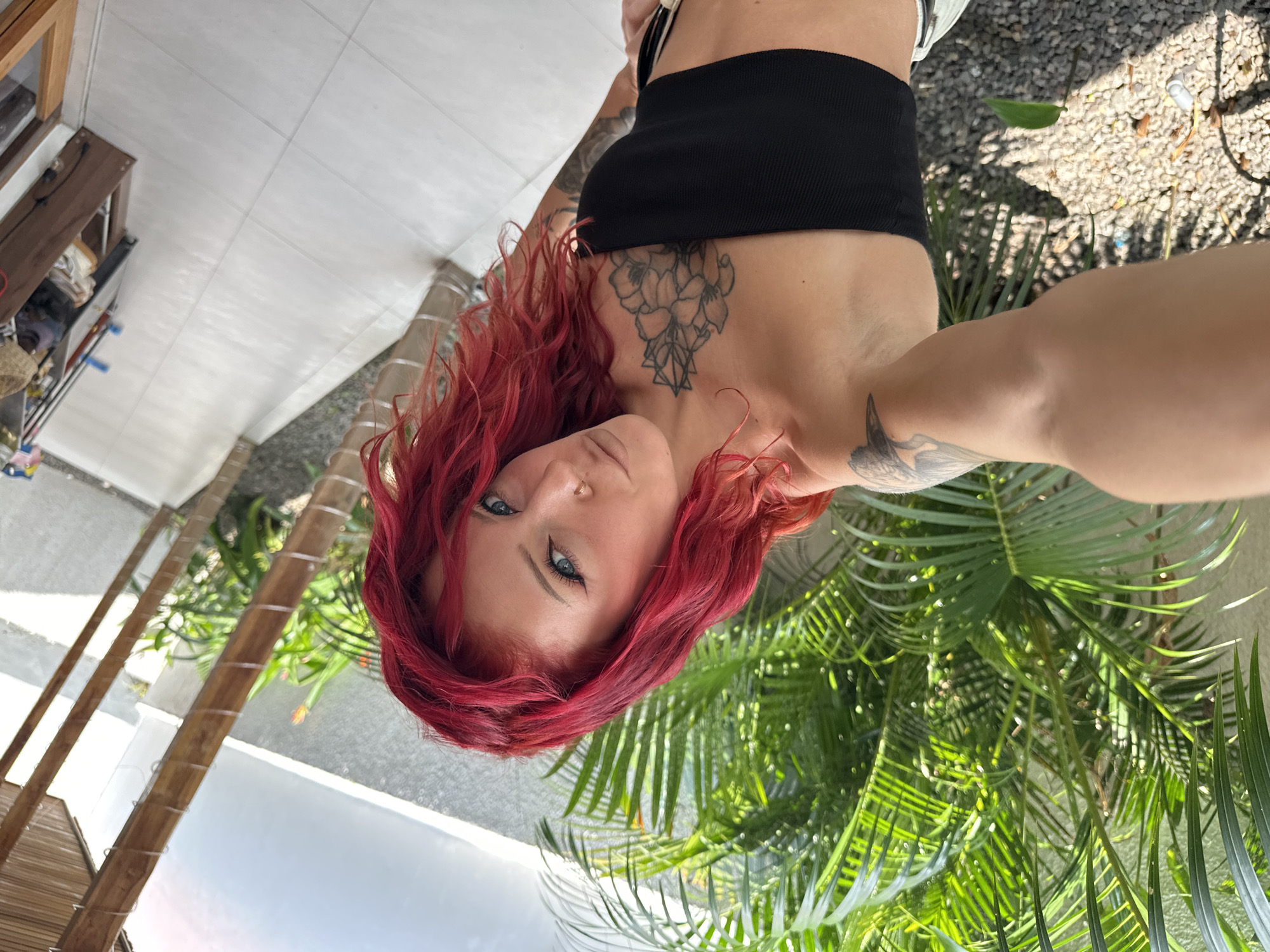 A woman with red, curly hair is in the frame. She has blue eyes and a tattoo on her chest of a flower or geometric design. She is wearing a black strapless top. Part of another tattoo is visible on her left arm. The woman is positioned in front of tropical plants with bright green leaves. Behind her, a tiled walkway and part of the building's exterior are visible. The location is suggestive of North Kuta, Indonesia, due to the lush, tropical vegetation. The image is taken from a tilted, close-up perspective.