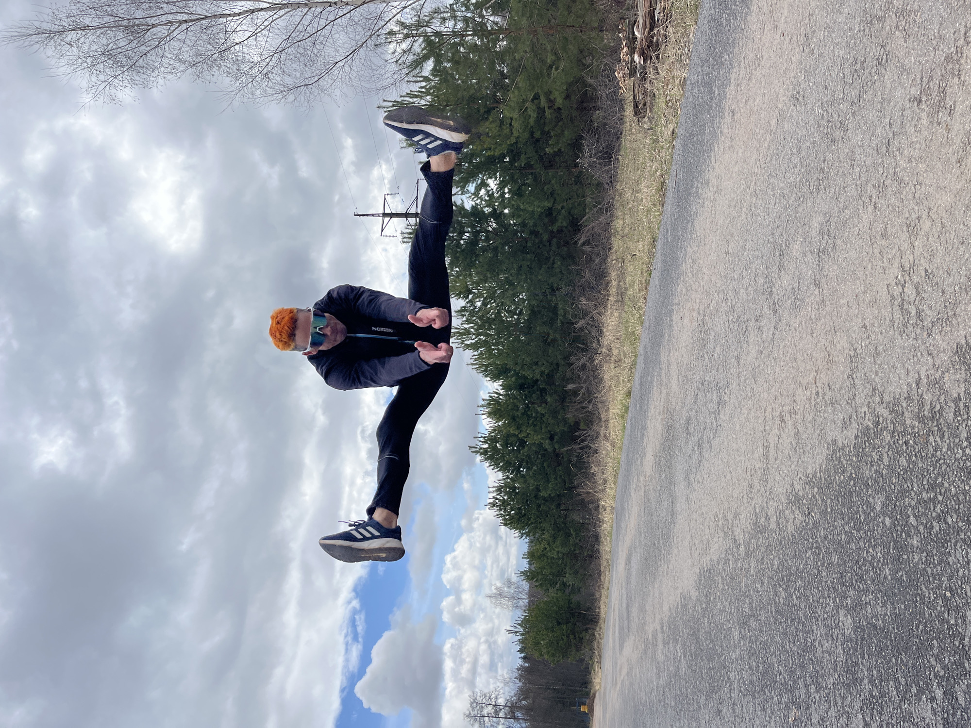 A person with orange hair, wearing black clothing and blue-and-white sneakers, is captured mid-air in a dynamic pose that might be a jump or a dance movement. The background shows a road on the right, trees, and a cloudy sky. A utility pole is also visible in the background. The photograph was taken on a road in the city of Chaikovsky, Russia.