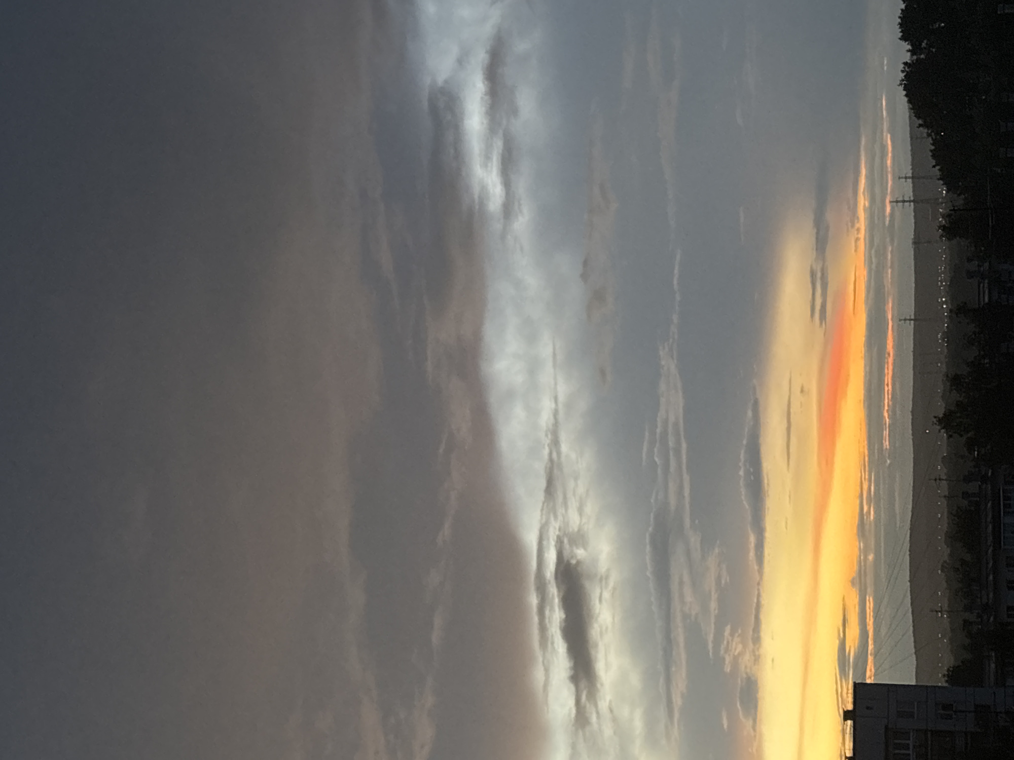 The image captures a dramatic sunset or sunrise over a town, likely Chaikovsky, Russia, viewed from an elevated perspective. The sky dominates the frame, showcasing a diverse range of cloud formations and striking colors.
In the upper portion of the sky, dark grey and dull white clouds stretch across, appearing heavy and diffused, with faint hints of sunlight filtering through in streaks. Below this, a more central band of clouds is illuminated with brighter, silvery-white tones, creating a sense of depth and ethereal light. Further down towards the horizon, the sky transitions into a vibrant display of warm colors. A prominent horizontal strip of brilliant orange and rich yellow light dominates this section, indicating the sun's low position. Patches of lighter, almost pinkish-orange hues are visible amidst the yellow and orange glow.
Along the horizon, a dark, undulating line suggests distant hills or mountains. Silhouetted against the colorful sky are several power lines and transmission towers, with faint, scattered lights suggesting a distant settlement or infrastructure. In the lower right portion of the frame, dark silhouettes of trees and buildings are visible, indicating the foreground of a town or city. The overall lighting is low, with strong backlighting from the sun creating dramatic contrasts between the illuminated clouds and the dark foreground elements. There is no visible text in the image.