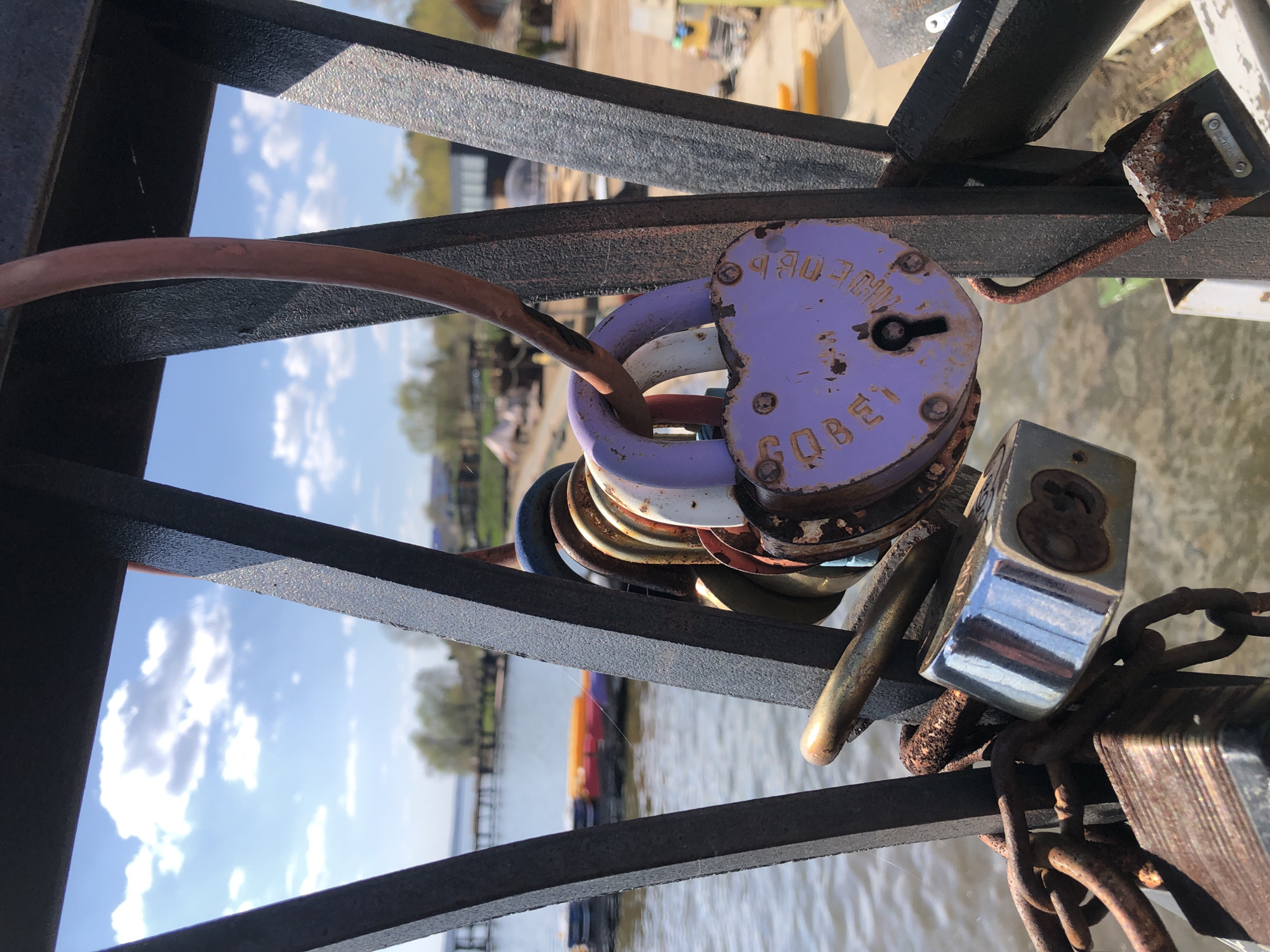 The image shows a close-up of multiple padlocks attached to a metal structure, likely a bridge railing, with a blurred background of water, trees, and buildings. The padlocks vary in size, shape, color, and material, with some showing signs of rust and wear. The most prominent padlock is heart-shaped and purple with the word "Любовь" (Love) written on it in Russian. The metal structure appears to be a framework of bars with a dark, possibly painted finish. The background indicates an outdoor setting, with a body of water visible, along with distant trees and structures suggesting an urban or semi-urban area. Based on the presence of the Russian word and the context of "love locks" being commonly attached to bridges, the location is likely a bridge in a Russian-speaking region, although the exact location in Votkinsk cannot be determined from the image alone.