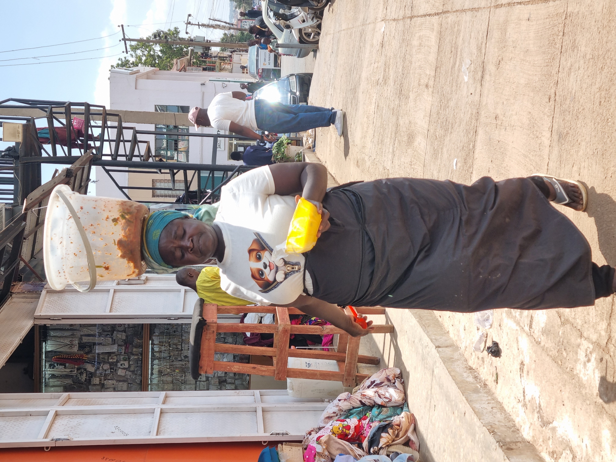 A woman stands on a paved street in Jos, Nigeria. She wears a white t-shirt with a puppy print, a black skirt, and sandals. On her head, she carries a large, stained white bucket. She holds a yellow plastic bag and a red object in her hands. To her left is a store with a metal security gate and a display case full of jewelry. A pile of textiles is on the ground. In the background, another man in a white shirt and blue jeans is walking on the same street, with parked vehicles and buildings visible further in the background. Power lines are overhead.