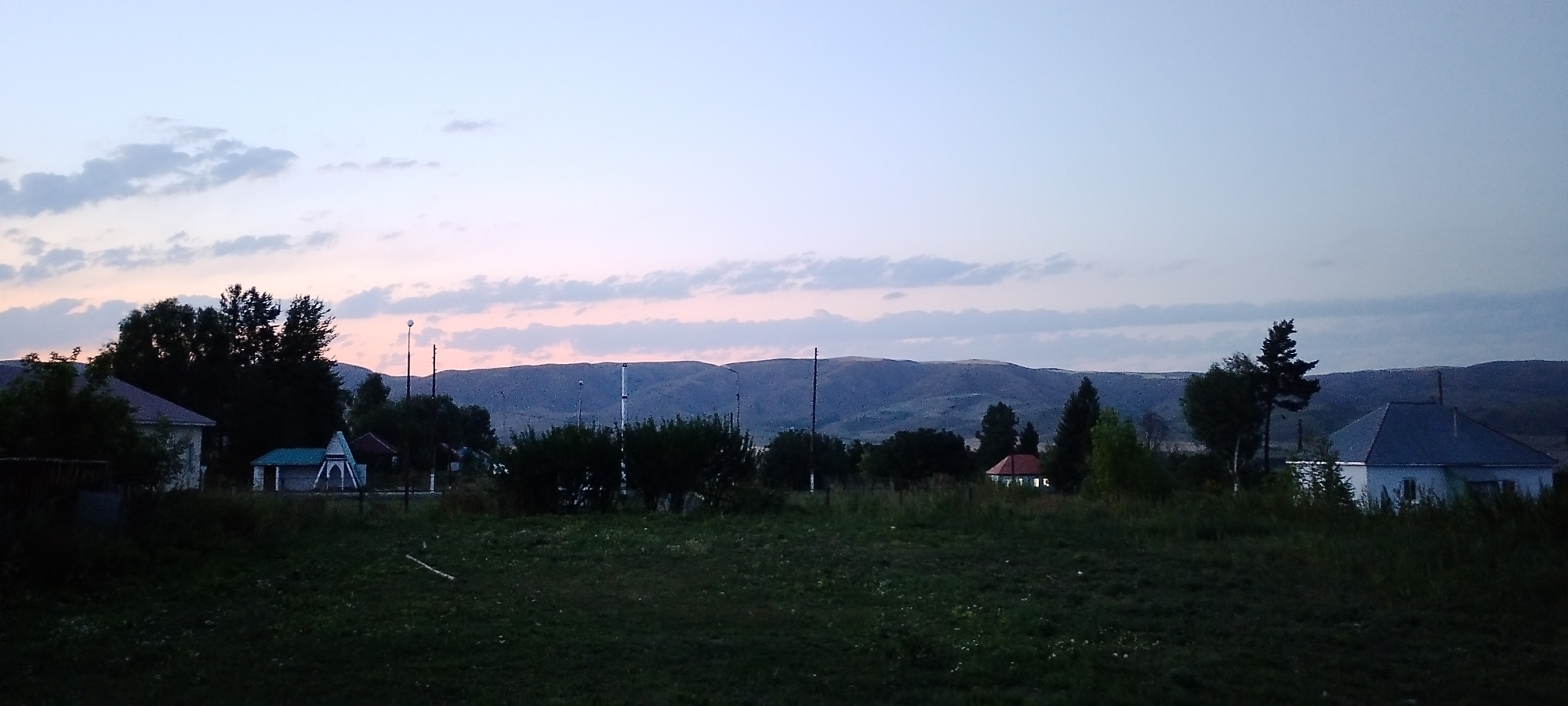 The image depicts a rural landscape at dusk, likely in the city of Lepsy, Kazakhstan. In the foreground, a dark, grassy field stretches across the frame. In the middle ground, several structures are visible. To the left, a white house with a dark roof is partially obscured by trees. Next to it, a smaller building with a turquoise roof and white walls features an arched entryway. Further to the right, another house with a red roof is illuminated from within. Scattered trees and some lampposts are also present. In the background, rolling hills are silhouetted against a pale sky that transitions from light blue at the top to soft pink and purple hues near the horizon, suggesting the setting or rising sun. Thin, wispy clouds are scattered across the sky. The overall impression is one of a quiet, twilight scene in a rural setting.