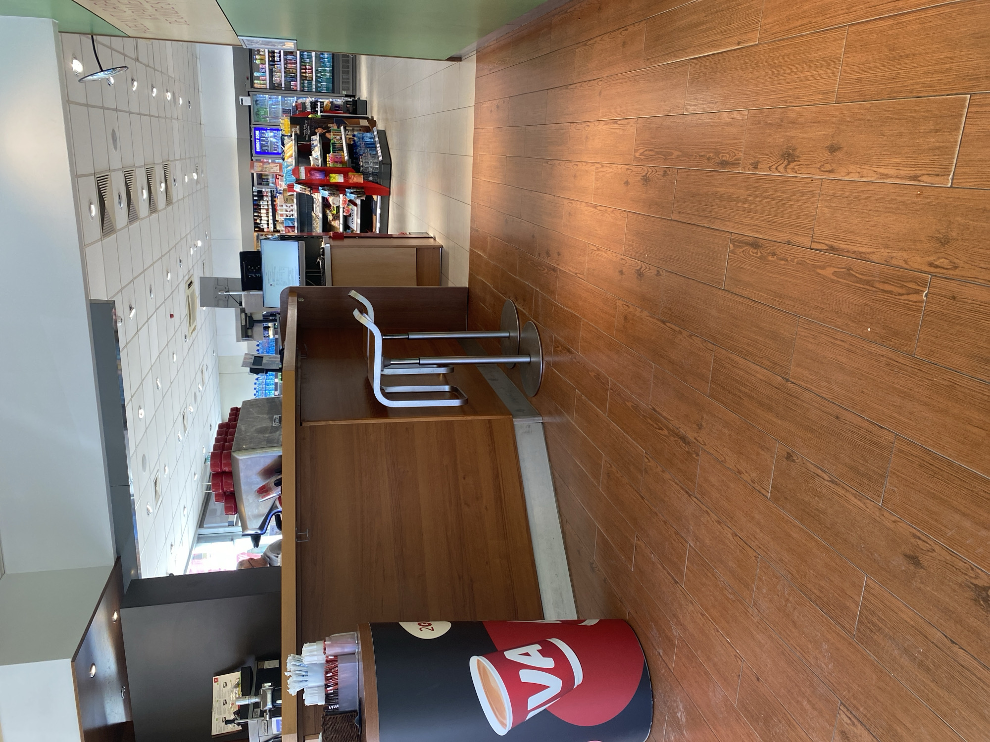 The image shows the interior of a commercial space, likely a cafe or convenience store, located in Varna, Bulgaria. The foreground features a wooden floor and a counter with two bar stools. A promotional display for VIVA coffee stands adjacent to the counter. In the background, shelves stocked with groceries and beverages are visible. The lighting is bright and artificial, suggesting an indoor environment. The scene is static, with no visible people or actions taking place at the moment.