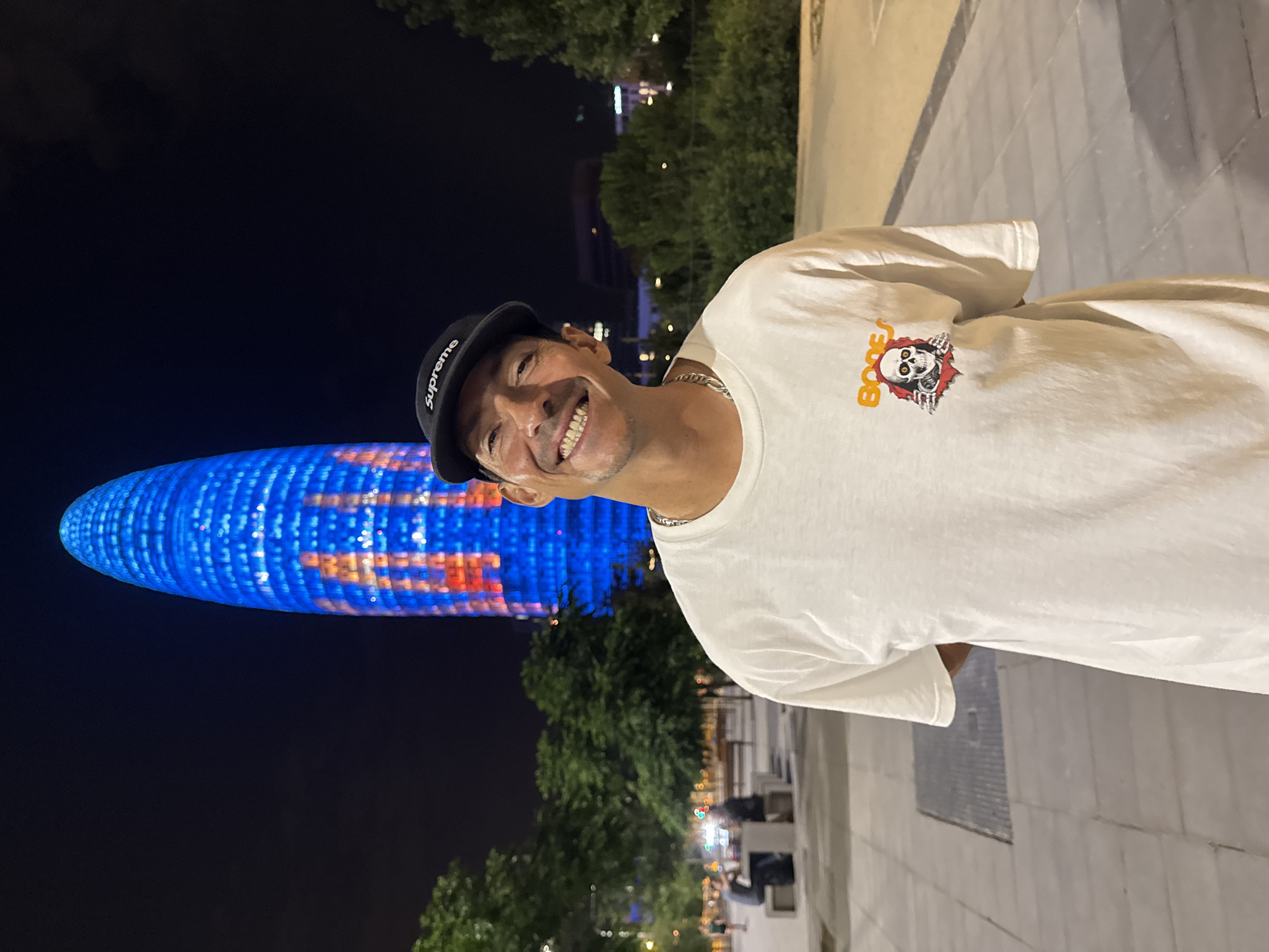 A man wearing a white t-shirt with a graphic of a skeleton and the word "Bones" on it, and a black baseball cap with "Supreme" embroidered on it, is smiling at the camera. He is standing in front of the Agbar Tower in Barcelona, Spain. The tower is illuminated with blue and red lights. In the background, trees and people can be seen. The ground is paved with concrete tiles. The sky is dark, indicating that it is nighttime.