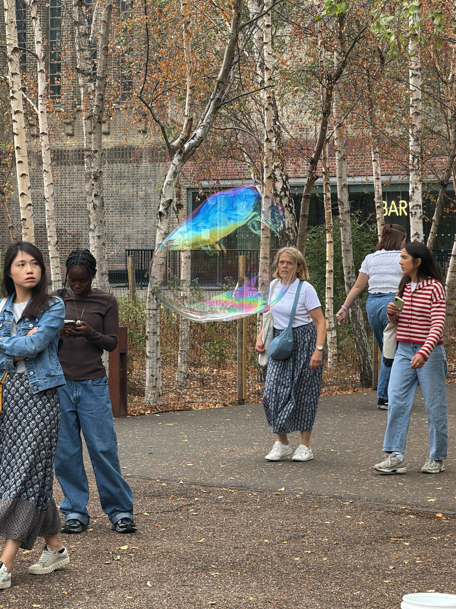 This is an outdoor scene in a park setting, possibly in London, United Kingdom, with a backdrop of birch trees and a brick building. In the foreground, several individuals are present. On the left, a young Asian woman in a denim jacket and patterned skirt stands with her arms crossed, looking to her left. Next to her, a Black woman in a brown long-sleeved shirt and blue jeans is looking down at her phone. A large, iridescent bubble drifts through the air between them and the center of the frame.
To the right of the bubble, a white woman with blonde hair, wearing a white t-shirt and a patterned blue and white skirt, is walking and looking forward. Further to the right, a woman with dark hair, wearing a white top and blue jeans, is visible in the background. Another young woman with dark hair, wearing a striped red and white cardigan and blue jeans, is in the right foreground, holding a phone. The ground is paved and scattered with fallen leaves. The overall atmosphere suggests a casual, possibly autumnal day in a public space.