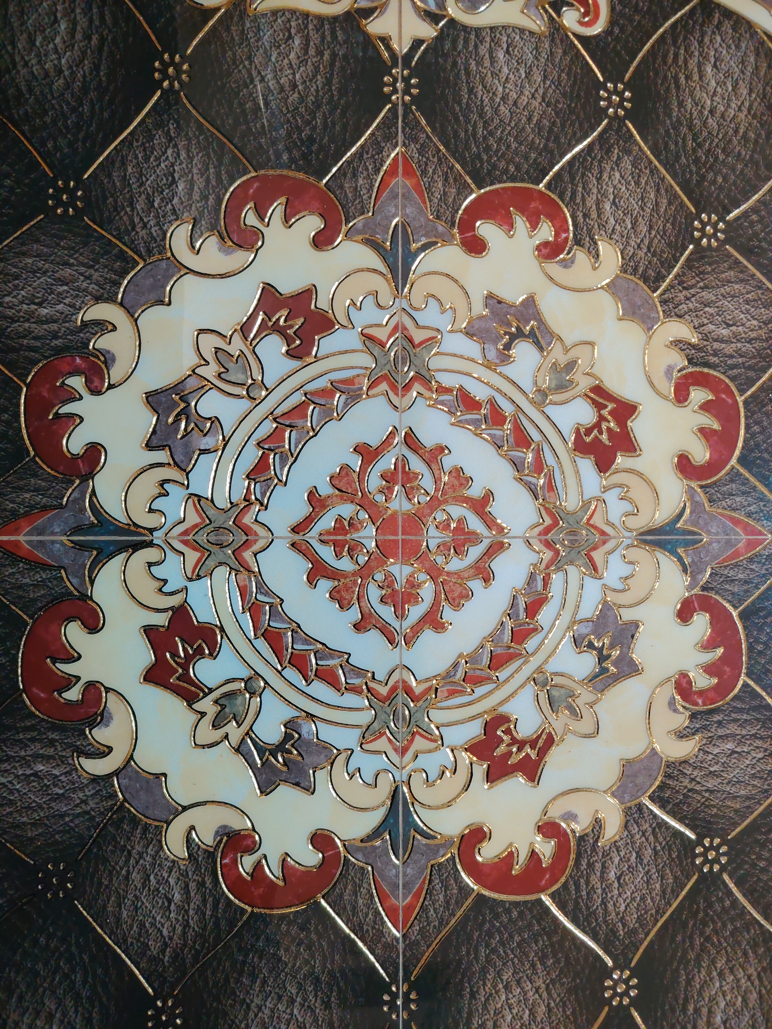 The image shows a close-up of a decorative tile with a circular, mandala-like design. The design features intricate floral and geometric patterns in shades of red, cream, and purple, outlined in gold. The background of the tile has a dark, textured appearance resembling leather with a diamond-stitched pattern and small gold embellishments. There are no people or animals present in the image. The location in Vnukovo, Russia, is not evident from the visual information provided in the image itself.