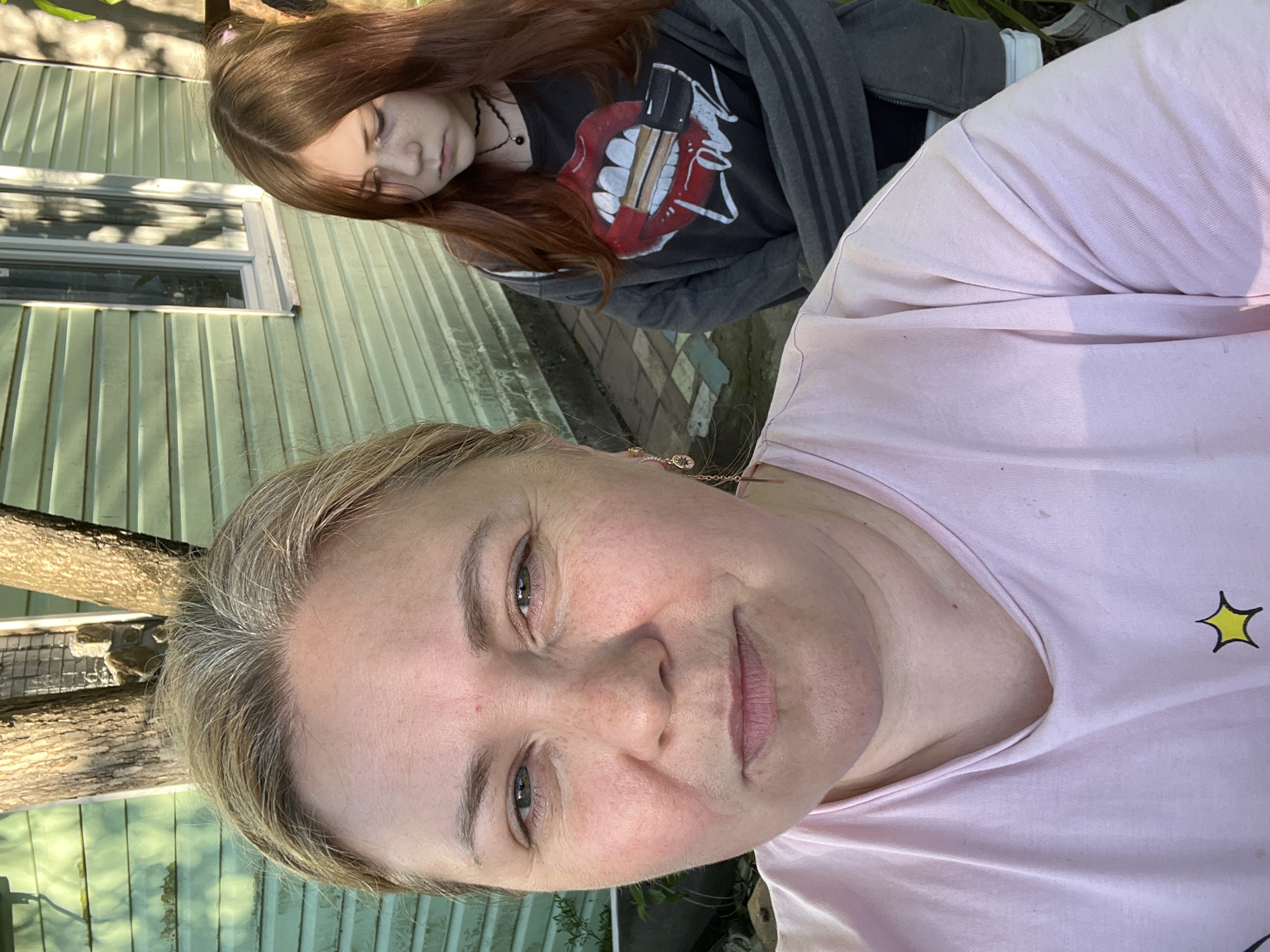 The image is a selfie of a fair-skinned woman with blonde hair. She is wearing a pink t-shirt with a yellow star. Behind her is a young woman with long auburn hair, a black choker necklace, and a black t-shirt with a red tongue and lipstick design under a gray hooded jacket. The background features a green building with a window and a tree. The location is Vesyoloye, Russia.