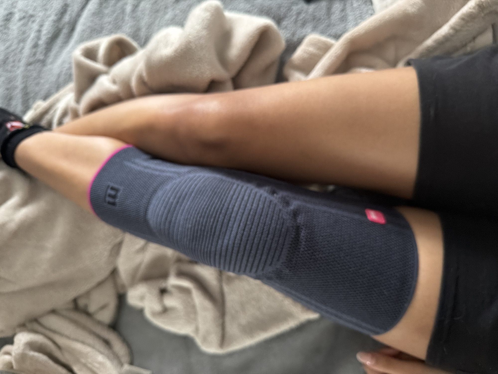 A close-up, high-angle shot shows a person's legs resting on a grey surface, possibly a bed or sofa, partially covered with a rumpled, beige-colored cloth. The person wears black socks and black shorts. The left knee has a dark blue knee brace with horizontal ribbed texture over the kneecap area and a pink band at the upper edge. The leg without the brace lies on top of the braced leg. The person's fingers are visible at the lower right. The location is not discernible from the image.