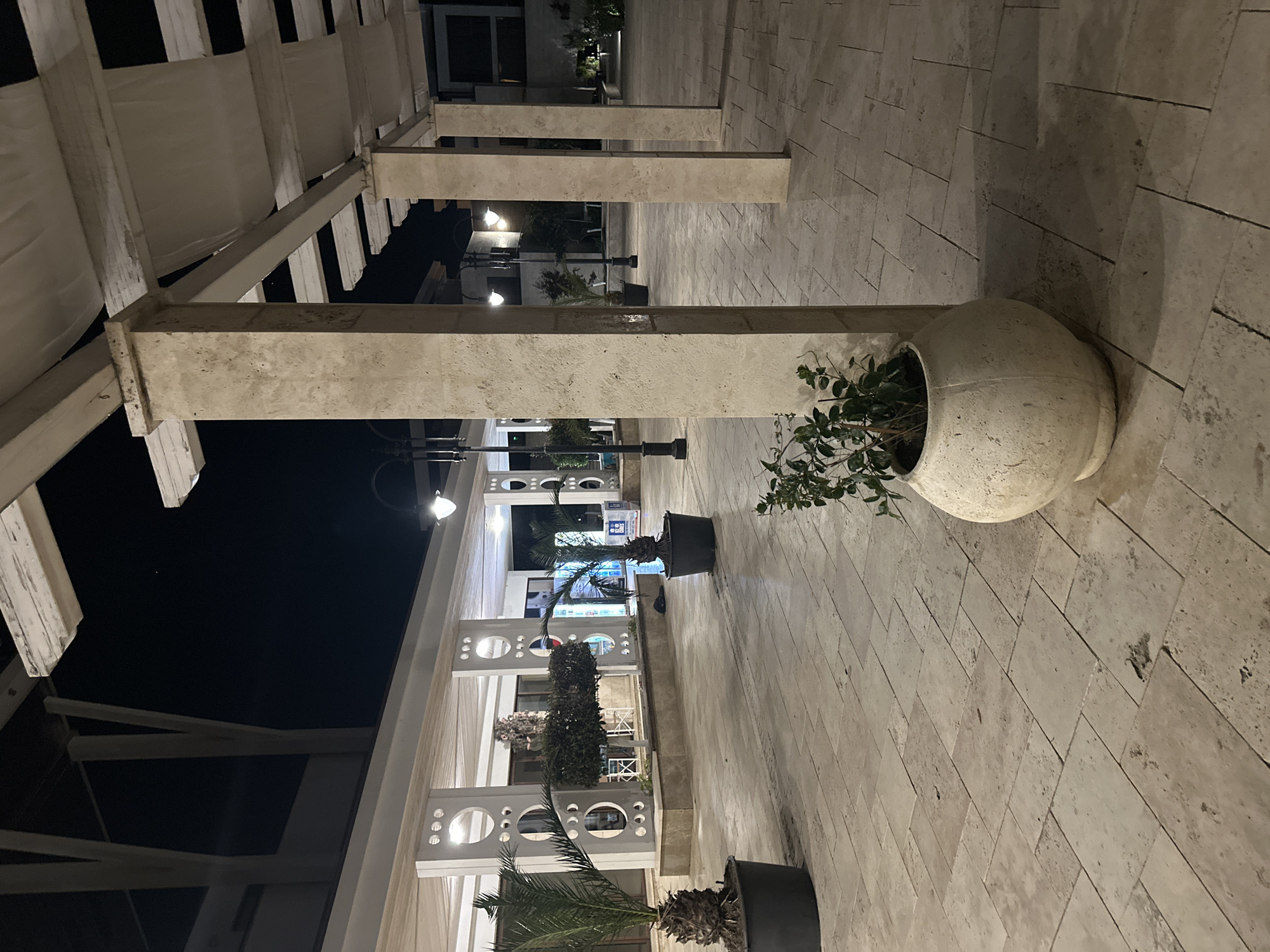 A nighttime exterior shot shows a paved walkway with large, light-colored stone tiles. A covered walkway with a white fabric roof supported by substantial stone pillars runs along the right side of the image. Several palm trees in large black pots are placed around the walkway, with one in the foreground on the left and another further back on the right. In the center background, there is a building with decorative circular patterns on its columns and a light-colored facade. The area is illuminated by streetlights and ambient light from the building. There are no people visible in the scene.