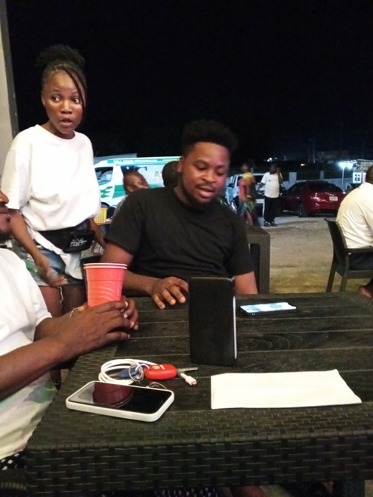 The image shows a group of people at an outdoor gathering in Lokoja, Nigeria, at night. A young woman with braided hair stands with a surprised expression, wearing a white t-shirt and shorts with a fanny pack. A man in a black t-shirt sits at a dark wicker table, seemingly engaged in conversation. Another person, partially visible, sits to the left, holding a red plastic cup.
On the table are a black wallet, a white smartphone, a set of keys with a red fob, a charger cable, and a white napkin. Other people are visible in the background, and there are cars parked nearby. A building with lit windows is in the distance. An ambulance is visible behind the woman.