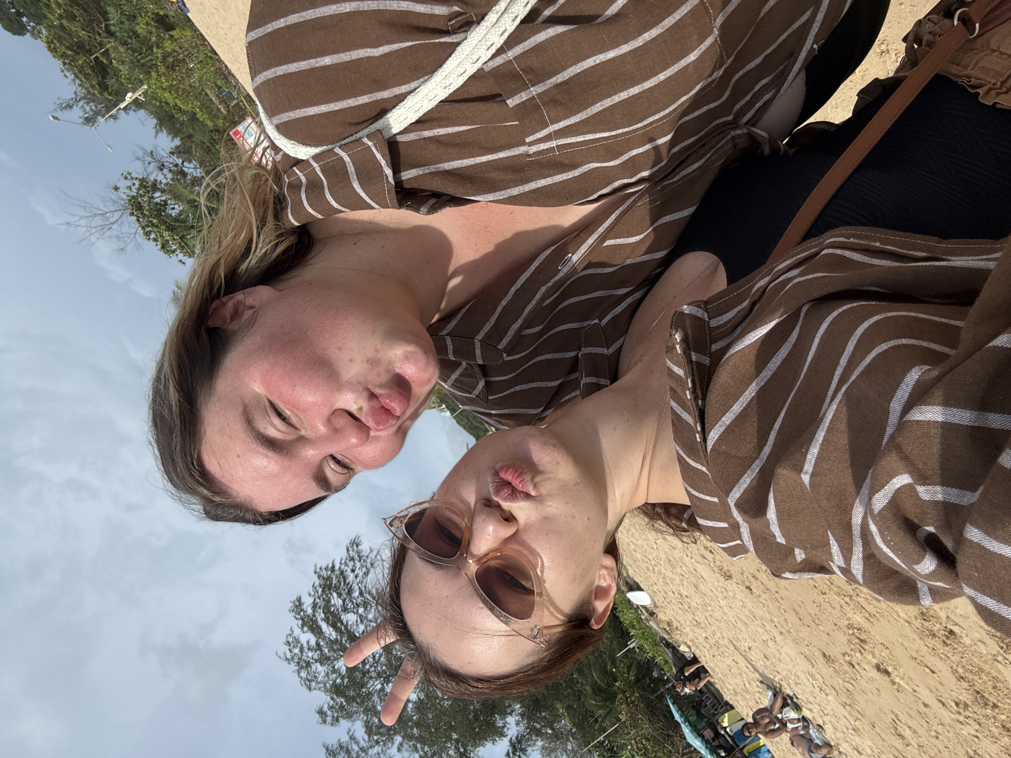 Two women are taking a selfie on a sandy beach in Rawai, Thailand. The woman in the foreground is wearing sunglasses and making a kissing face with bunny ears formed by her fingers. The woman behind her is also making a kissing face. Both women are wearing brown and white striped shirts. In the background, there are trees, beach umbrellas, and other people relaxing on the sand. The sky is cloudy.