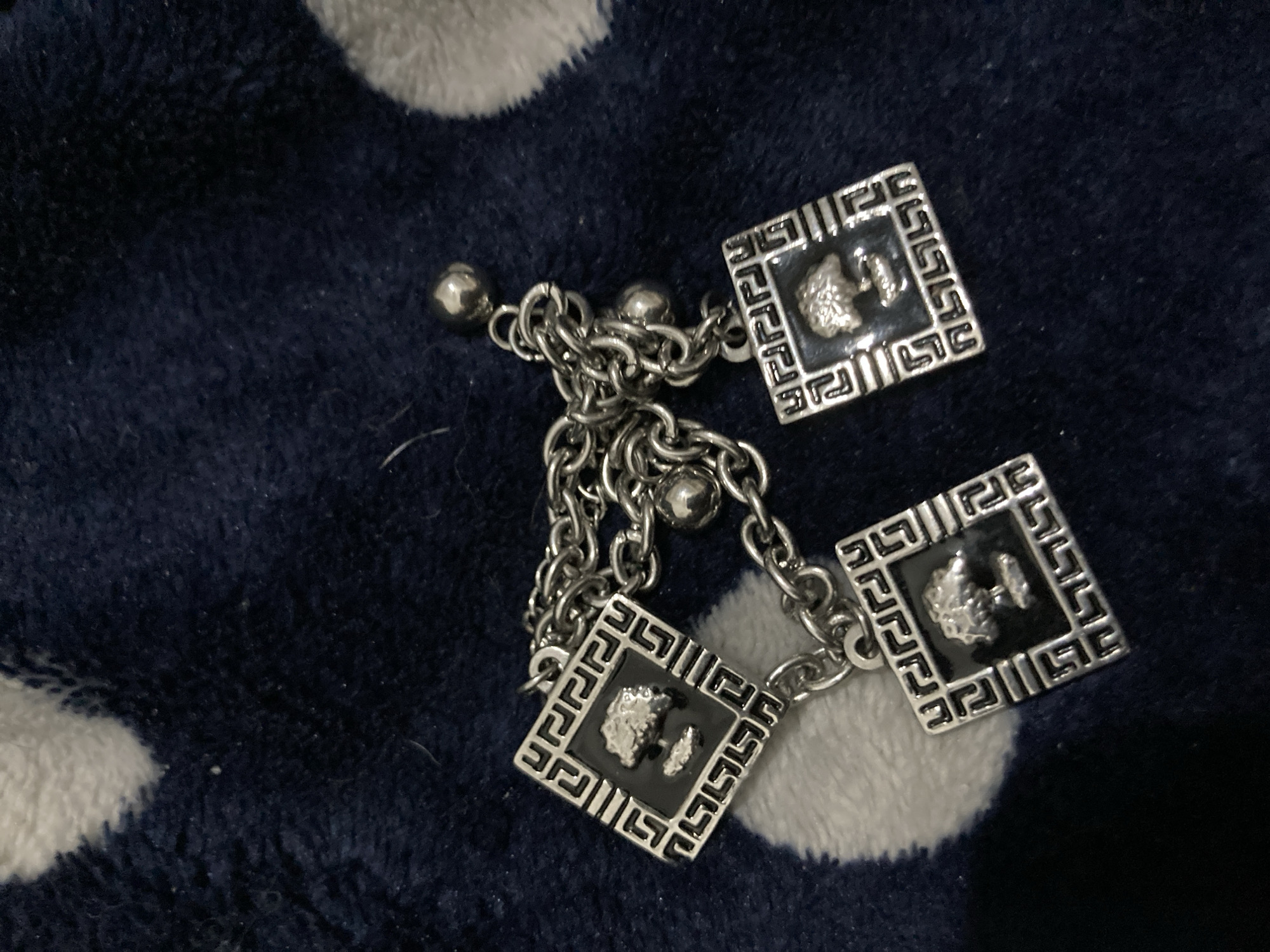 A silver chain bracelet with three square Versace charms and two silver balls lies on a dark blue blanket with large white polka dots. The charms are silver with a black background and a raised silver Versace Medusa head design. The bracelet is in the center of the frame. The location is unknown.