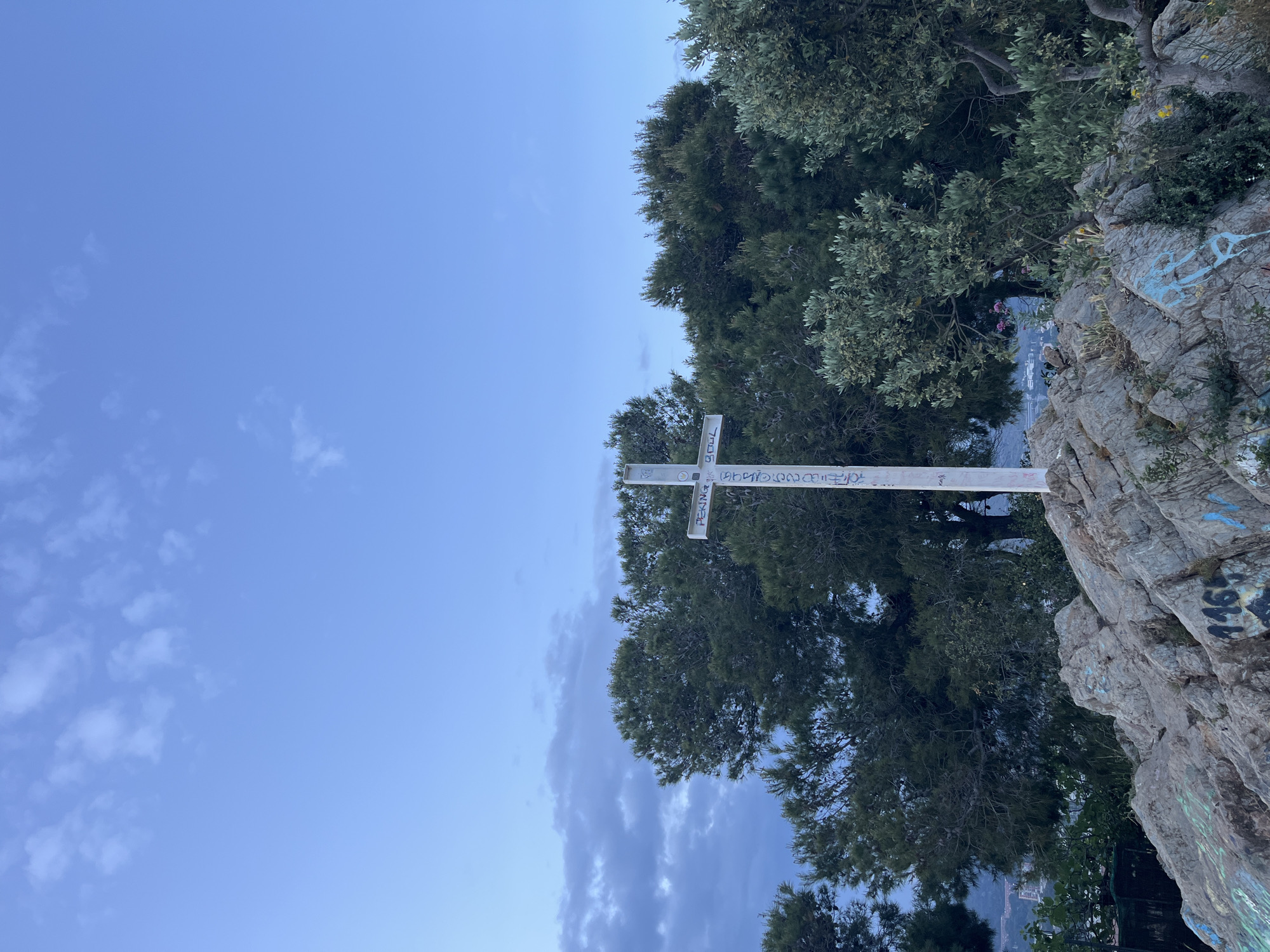 The image shows a white cross situated on a rocky outcrop in Barcelona, Spain. The cross is covered in graffiti, including the word "PEKING." The rock face also has graffiti. Trees surround the cross and the rock. The sky is blue with some light clouds.