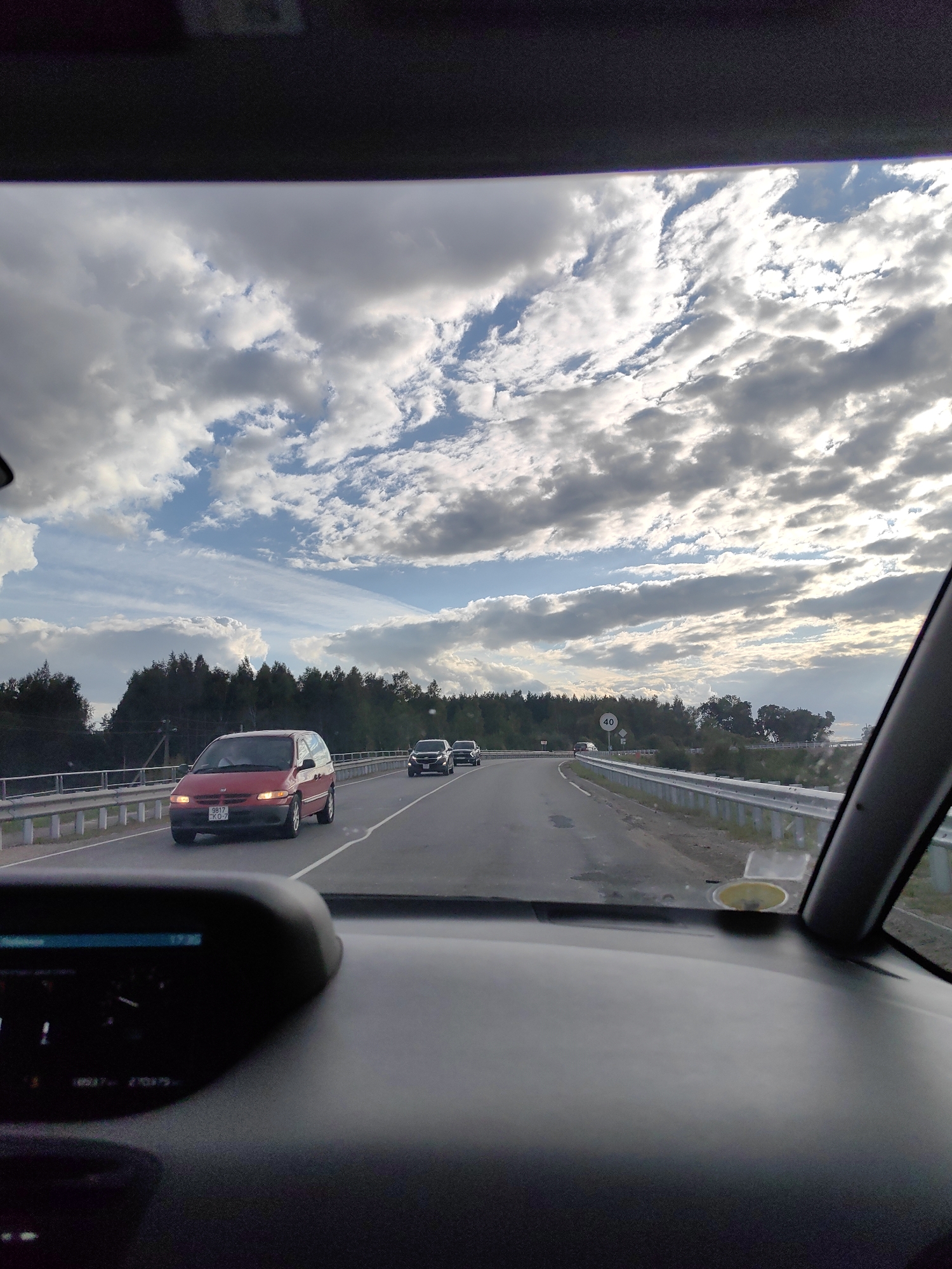 The image shows a view from inside a vehicle traveling on a highway. The road appears to be in a rural area with trees lining the sides. There are several cars on the road, including a red minivan and two darker sedans behind it. A speed limit sign indicating "40" is visible on the right side of the road. The sky is filled with dramatic clouds, suggesting either early morning or late afternoon lighting. The dashboard of the vehicle is partially visible in the foreground, with some instruments showing. The image quality is grainy, indicating it was likely taken with a mobile phone camera. The license plate of the red minivan is visible, reading "9817 KO-7".
