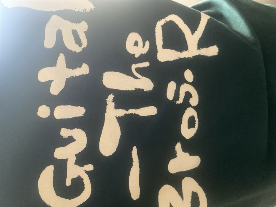 The image displays a close-up view of a dark teal or dark green textile surface featuring bold white lettering with irregular, brush-stroke-like edges. The text is arranged in three visible lines: the top line reads "Guital", the middle line shows a horizontal dash followed by "The", and the bottom line displays "Bros. R". A bright, diffuse light source is evident in the upper right portion of the frame, causing a glare across that area of the fabric. No individuals, additional objects, specific actions, or environmental context are discernible within the image. The location in the city of Katcha, Nigeria, cannot be determined from the visual information presented.