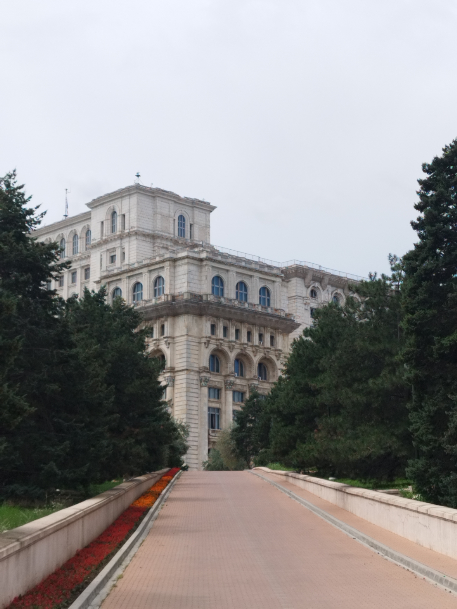 The image shows a frontal view of the Palace of the Parliament in Bucharest, Romania, under a gray, overcast sky. The imposing white stone building dominates the upper half of the frame, featuring multiple levels, arched windows, and ornate architectural details. Tall, dark green evergreen trees flank the building on both the left and right sides, partially obscuring the lower sections of the structure. In the foreground, a wide, paved walkway or road made of reddish-brown bricks leads upwards towards the palace. Along the left side of this walkway, a raised, light-colored concrete barrier borders a long, narrow flower bed filled with vibrant red and orange flowers. The overall scene conveys a sense of grandeur and scale associated with the monumental architecture of the palace.