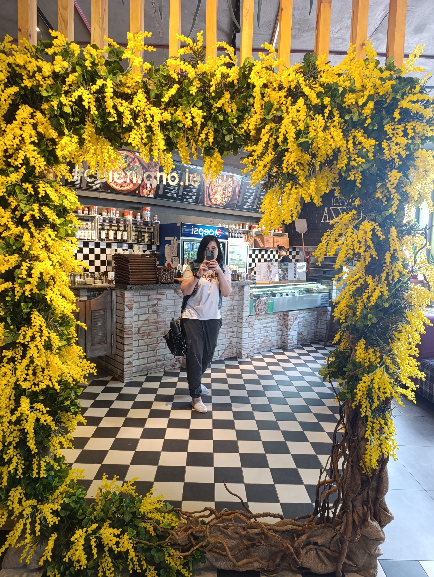 This is a full-shot, eye-level, indoor photograph taken inside a restaurant in Bila Tserkva, Ukraine. A woman with dark hair, wearing a white t-shirt, dark pants, and white shoes, is standing in the center of the frame, taking a selfie with her phone. She has a black backpack slung over her right shoulder. The restaurant has a distinctive black and white checkered floor. The foreground is dominated by a large, arched floral decoration featuring numerous yellow blossoms and green leaves, supported by rough, exposed tree roots wrapped in burlap. In the background, a counter area is visible with brick and checkered tile walls, shelves stocked with food items, and a glass display case. Menus are displayed on the wall behind the counter, and a large blue refrigerator is visible to the right. The overall scene suggests a vibrant and perhaps festive atmosphere within the establishment.