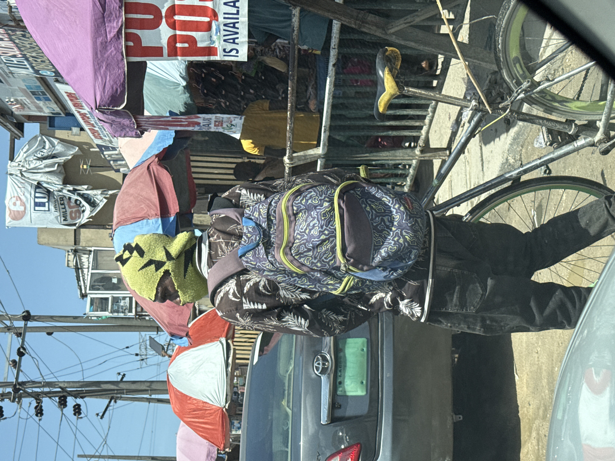 A man wearing a patterned shirt and a yellow knit cap with black geometric designs carries a patterned backpack on a bicycle in Lagos, Nigeria. The man is surrounded by colorful umbrellas and various signs, with a gray Toyota car partially visible. Overhead, a network of power lines stretches across a clear blue sky.