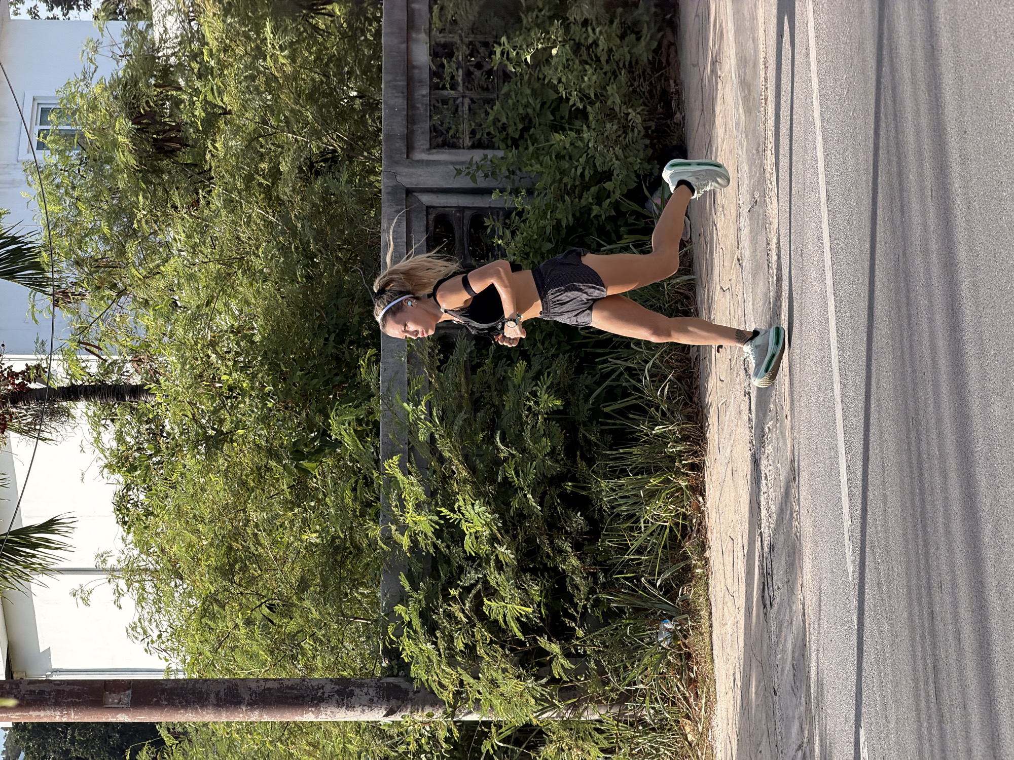 An adult female with blonde hair in a ponytail, wearing a black sports bra, black patterned shorts, and white and green running shoes, is captured in mid-stride, running on a paved sidewalk alongside a road. She has a smartwatch on her left wrist and is wearing earbuds. The background features lush green foliage, a concrete fence, and a portion of a white building. The scene appears to be outdoors in a tropical or subtropical environment, consistent with the location of Ko Samui District, Thailand, indicated by the dense vegetation and the style of the building and fence. The lighting suggests it is daytime, likely morning or afternoon, with shadows cast on the road.