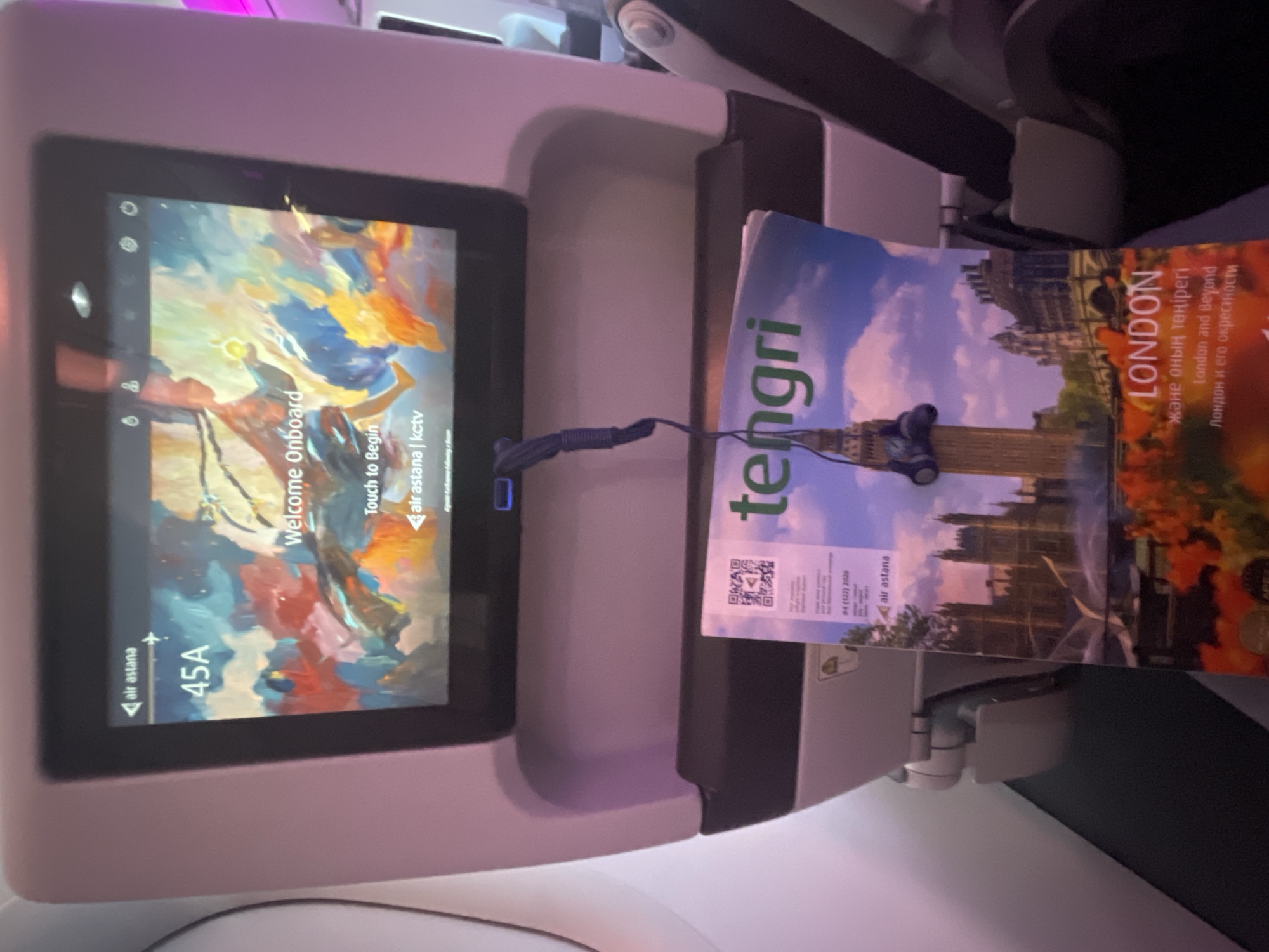 An in-flight entertainment screen with an abstract painting and "Welcome Onboard" text is displayed. Below the screen, a magazine titled "Tengri" with images of London is visible, with a pair of earphones attached. The scene appears to be inside an airplane cabin, with part of a seat and armrest visible. The location is identified as Arnavutköy, Turkey.