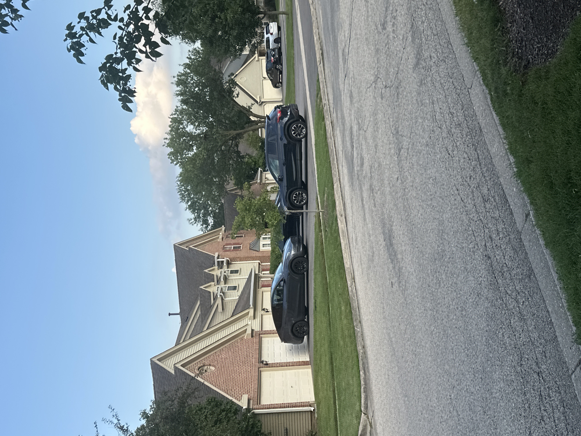The image shows a suburban street scene in Schaumburg, United States of America, on a clear day with a few clouds. In the foreground, a gray asphalt road is visible with a concrete curb and green grass. Several houses with pitched roofs and attached garages line the street. In front of one house, two cars are parked: a dark gray Tesla Model Y and a black Mazda CX-5. Further down the street, more cars are visible parked in front of other houses, including a white SUV and a dark-colored sedan. Trees and foliage are present throughout the scene, adding to the suburban atmosphere.