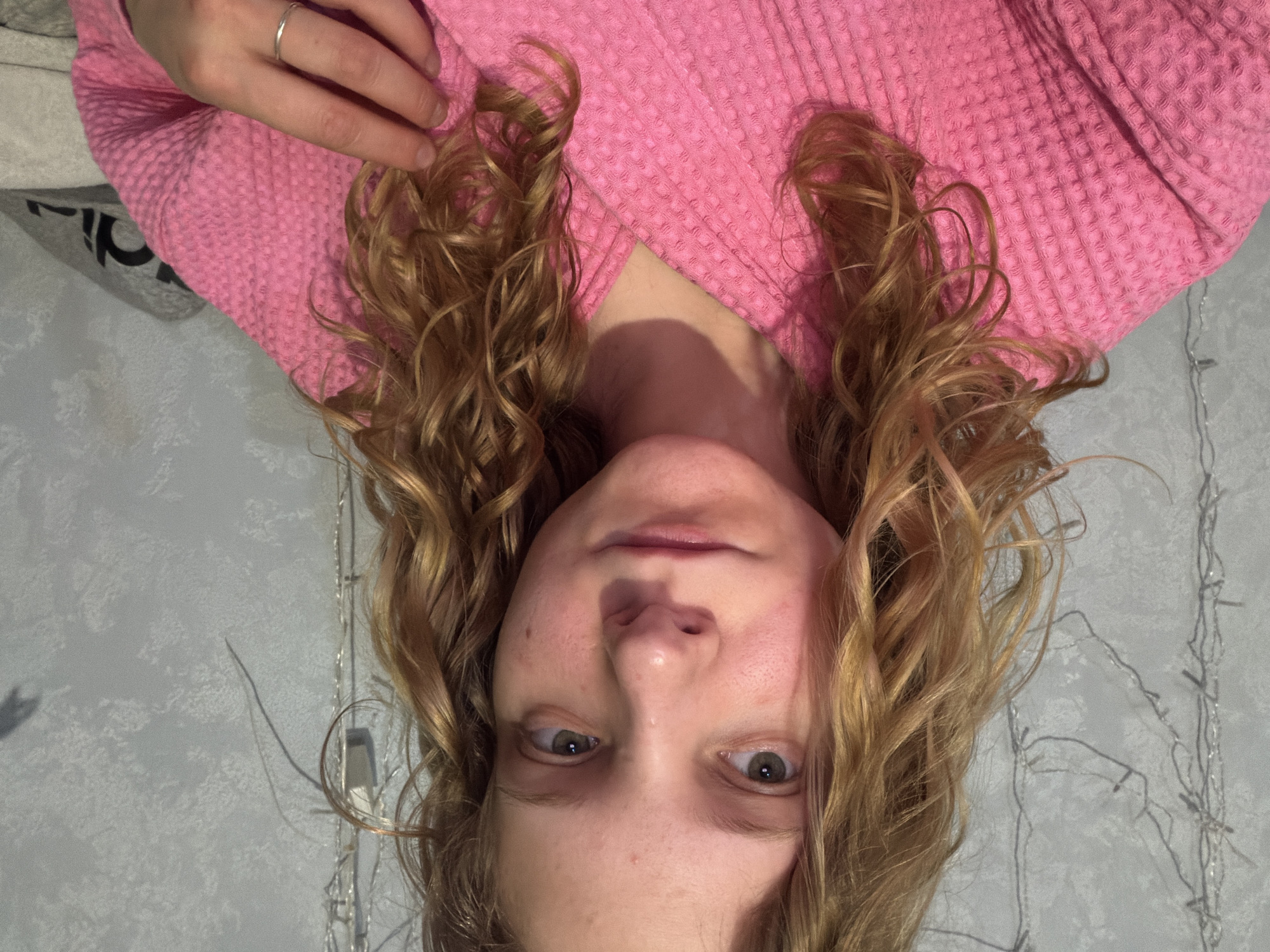The image depicts a person with light skin and curly blonde hair, wearing a pink waffle-textured robe or shirt. The person's face is visible, with light-colored eyes and a neutral expression. One hand is visible, with a silver ring on a finger. The background is a gray wall with a pattern resembling marble, and some string lights are visible along the edge of the wall. There is no information in the image to suggest it was taken in the city of Chaikovsky, Russia. The image is rotated upside down.