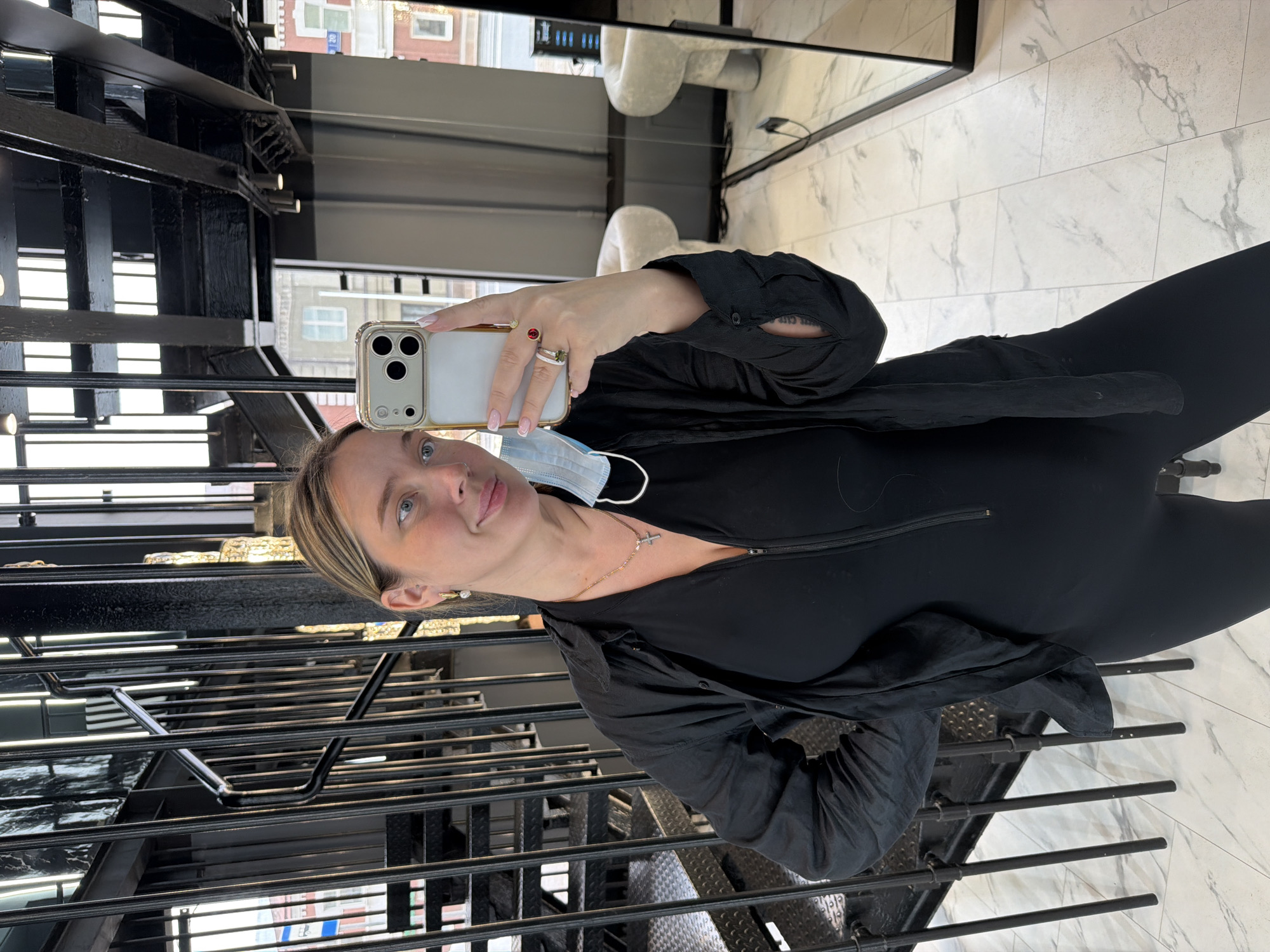 A woman stands on a black metal staircase, taking a selfie in front of a mirror. She is wearing a black jumpsuit, a black blouse, and a light blue surgical mask hanging on her neck. She has blonde hair pulled back, fair skin, and is wearing jewelry including rings and a cross necklace. The mirror reflects a modern interior with white marble-like floors and white furniture. The reflection shows the setting to be a building with a window showing a city street outside. Based on architectural styles observed, the location is suspected to be Moscow, Russia.