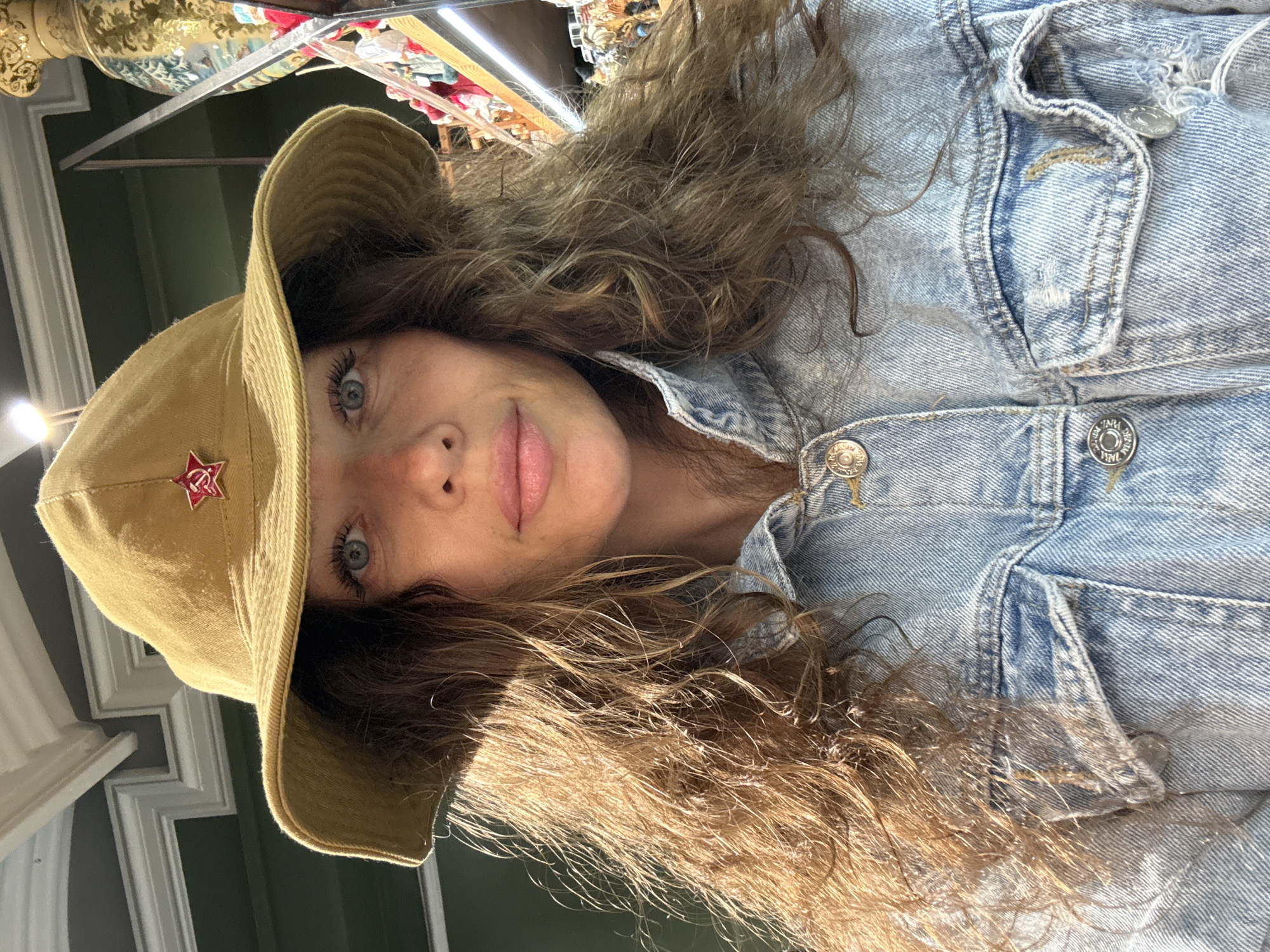 A woman with long, curly brown hair is taking a selfie in what appears to be a shop. She is wearing a tan bucket hat with a red star emblem and a light blue denim jacket. The background is out of focus, but shelves with various items, possibly souvenirs or decorative objects, are visible. The lighting suggests an indoor setting. The woman's eyes are blue and she has natural-looking makeup. She is looking directly at the camera.