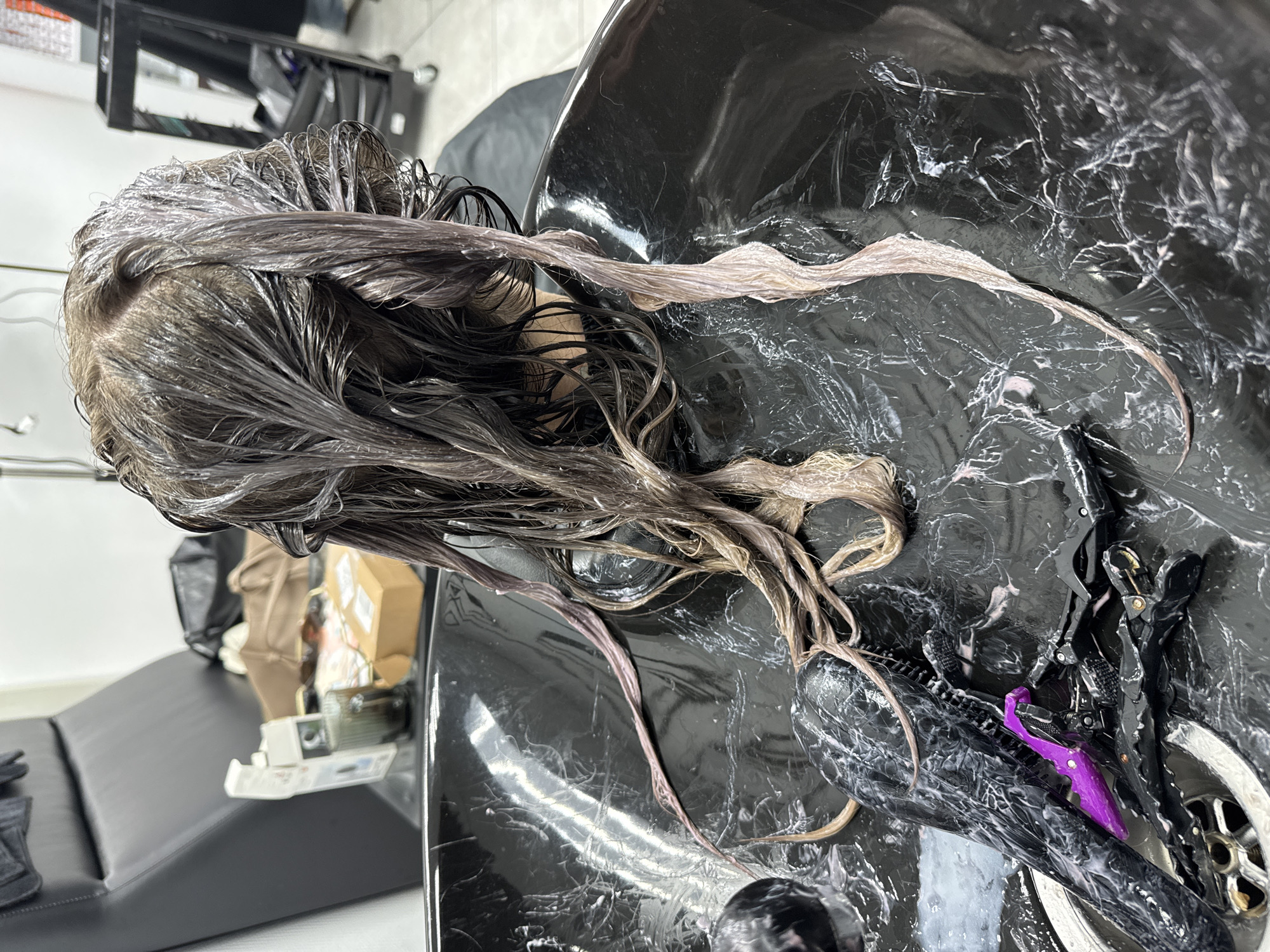 The image depicts a close-up view of a person's wet hair in a salon sink. The hair appears to be in the process of being colored or treated, with a light-colored product visible on the strands. Several black and purple hair clips are scattered in the sink basin, along with a black brush. The sink itself is black and has a marbled pattern. In the background, a black salon chair is visible, as well as some supplies and a cart. The scene is indoors, consistent with a hair salon environment. There is no visible indication of the location being in Chaikovsky, Russia.