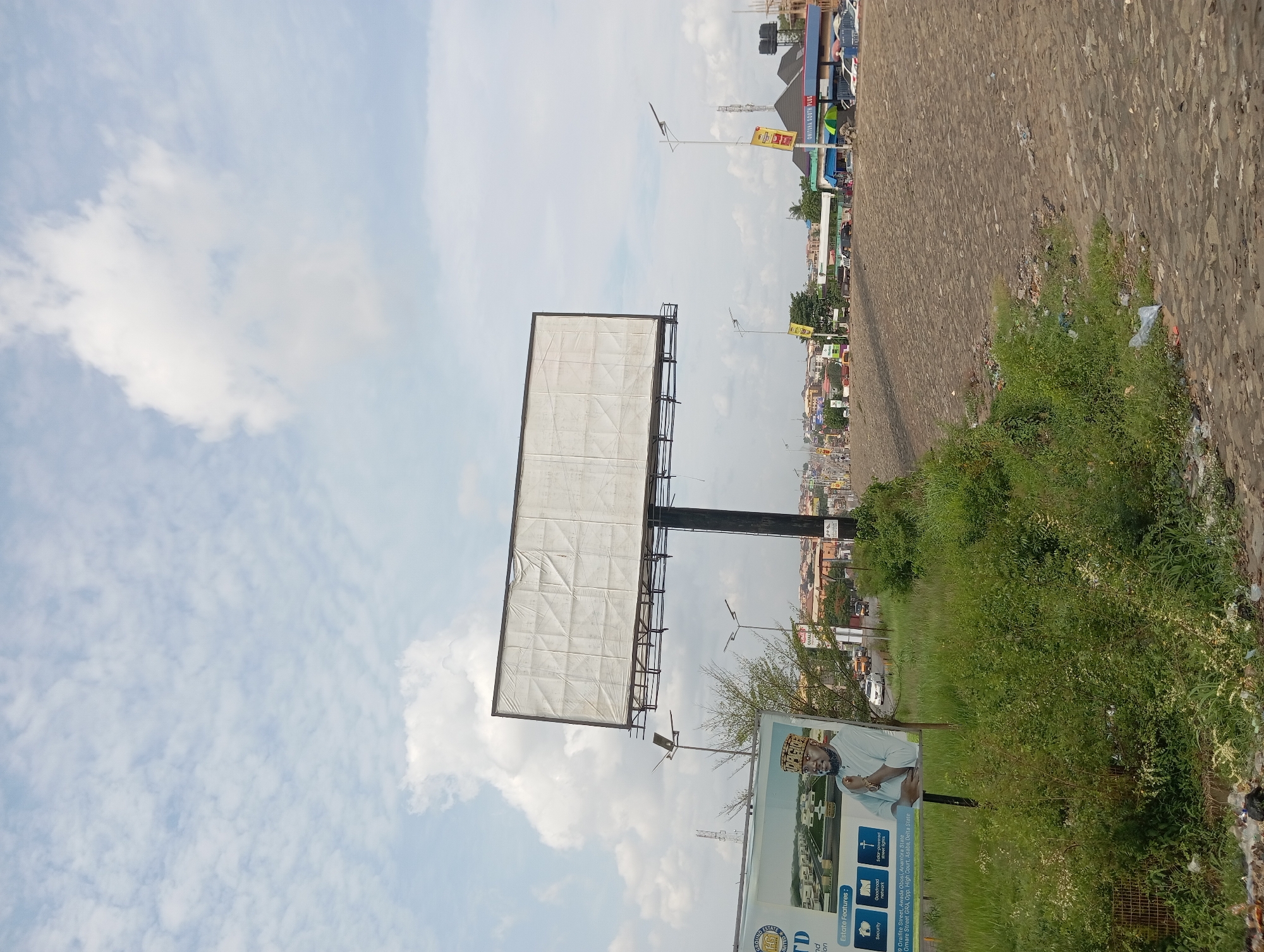 The image shows a scene in Nkpor, Nigeria. A large, blank billboard dominates the left side of the frame, partially obscured by clouds. In the foreground, a smaller sign displays an advertisement for real estate, featuring an image of a man and listing amenities. To the right, the roadside is lined with dense greenery and some litter. Beyond the vegetation is a rough, cracked road surface. In the distance, the cityscape is visible, with buildings of varying heights. Above, the sky is a mix of blue and white clouds.