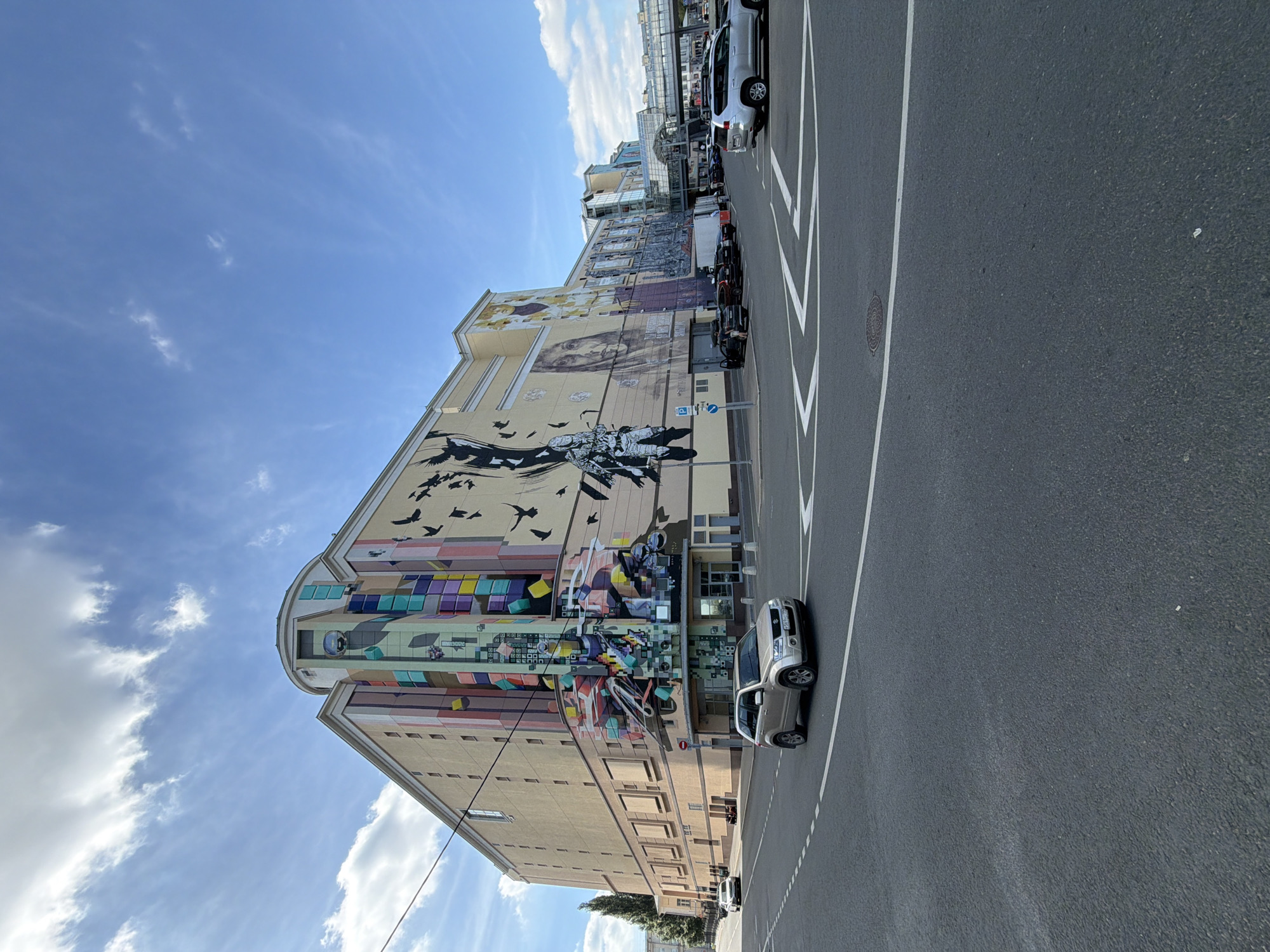 A large building with a vibrant mural on its side is the central subject of this image. The mural depicts a scene of birds in flight over abstract geometric shapes and what appears to be a stylized cityscape. On the street in front of the building, several cars are parked, including a silver SUV. The setting is urban, with the building occupying a significant portion of the frame. The time of day appears to be daytime, indicated by the bright sky with scattered clouds. The weather seems pleasant. The overall mood is neutral, focusing on the urban landscape and the artwork. There is a blue and white parking sign visible on the building. The image is oriented vertically, suggesting it was taken from a low angle or with the camera tilted. The building itself is multi-story and has a contemporary architectural style. The street in front of it is paved and marked with white lines, indicative of a parking area or traffic lane. The mural is extensive, covering a large section of the building's facade, and features a combination of black, white, and colorful elements.