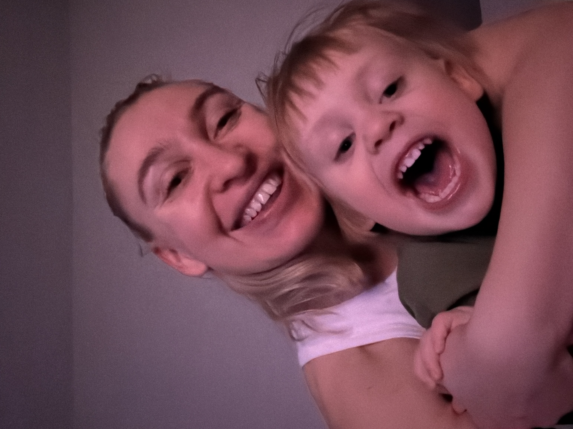 The image portrays an adult female embracing a young child in an indoor setting. The background consists of a plain, light-colored wall, illuminated by a warm, reddish-purple light source. The adult female, positioned behind the child, has light-colored hair and is smiling broadly, revealing both upper and lower teeth. She is wearing a white sleeveless top. The child, positioned in front, has light-colored hair with bangs and is depicted with an open mouth, appearing to laugh or shout, also revealing upper and lower teeth. The child wears an olive green top, and one of the child's hands is visible, clutching the adult's arm. Both individuals are looking directly towards the camera. The depicted scene is located in the city of Chaikovsky, Russia.