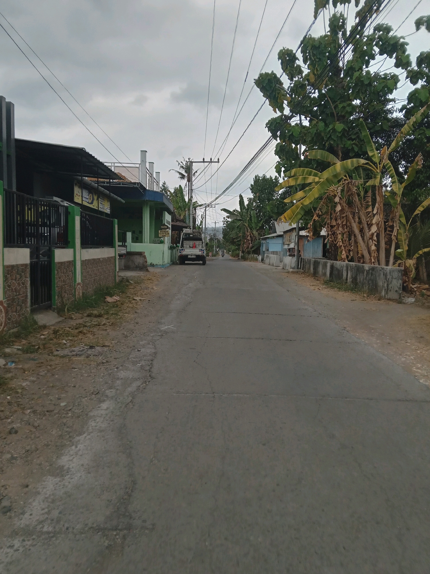 The image depicts a residential street in Banguntapan, Indonesia, under an overcast sky. The scene is viewed from the perspective of someone standing on the asphalt road. On the left side of the road, a low wall constructed of stone and concrete borders a property with a black metal fence. Beyond the fence, a building with a dark awning and green trim is visible. Several utility poles with a complex network of wires are present throughout the scene. On the right side of the road, a concrete retaining wall separates the asphalt from a dirt path, behind which are small houses and banana trees. A white pickup truck is parked on the right side of the road further down. The overall impression is of a quiet, somewhat rural-feeling street with typical infrastructure and vegetation for the region.
