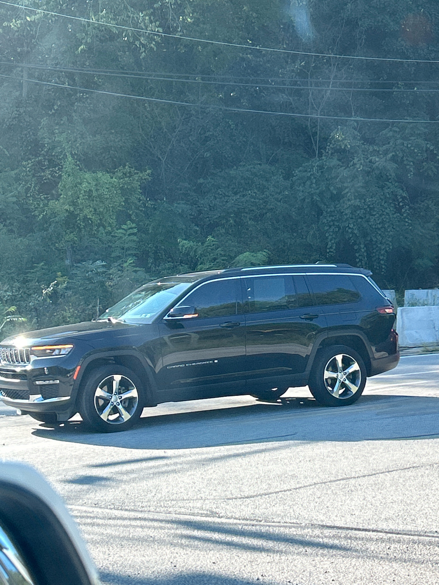 The image shows a black Jeep Grand Cherokee driving on a road with trees and foliage in the background. The sun is shining, casting shadows on the road. There are power lines visible above the trees. The car is in motion, and its headlights are on. There are no people visible in the image. The car appears to be in a semi-rural or suburban area, possibly on the outskirts of a city.