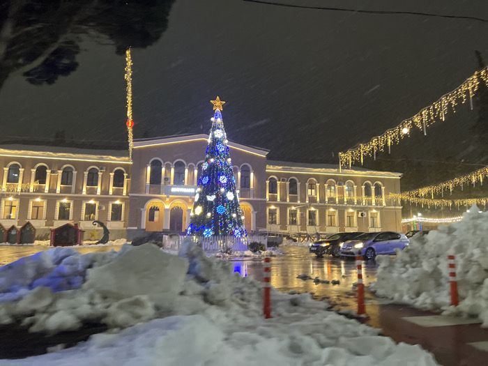 The image displays a nighttime winter scene in the city of Terjola, Georgia. Light snow is actively falling. A large, brightly illuminated Christmas tree, adorned primarily with blue and white lights and topped with a yellow star, stands centrally in the midground. Behind the tree is a two-story building, likely the Terjola Municipality, distinguished by multiple arched windows on its upper floor and rectangular windows on the lower. The building's architectural features are outlined with warm white string lights. A sign in Georgian script, "ტერჯოლა" (Terjola), is visible above the central entrance. Overhead, several strands of warm-toned, icicle-style string lights are suspended across the frame.
The ground is wet and reflective, indicating recent precipitation or melted snow. Large, uneven piles of white snow are prominent in the foreground and along the right side of the image. Partially visible red and white striped bollards protrude from the snow in the immediate foreground. Two passenger vehicles, a dark-colored sedan and a lighter-colored hatchback, are parked on the wet pavement to the right of the Christmas tree, their headlights reflecting on the surface. No individuals are visible in the scene.