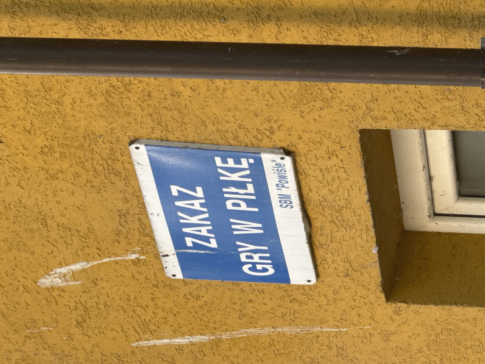 This image presents a close-up view of a textured, ochre-yellow building wall, illuminated by what appears to be bright daylight. A dark grey pipe, possibly a drainpipe, runs horizontally across the upper portion of the wall, casting a faint shadow below it.
The central subject is a rectangular blue and white sign, affixed to the wall and tilted slightly downwards to the left. The sign exhibits some wear, with visible scuffs and fading on its surface. White text on a blue background dominates the sign, reading:
"ZAKAZ"
"GRY W PIŁKĘ"
Below this, in smaller text, it states: "SBM "Powisle"".
Translated from Polish, "ZAKAZ GRY W PIŁKĘ" means "NO BALL GAMES" or "PROHIBITION OF PLAYING BALL". "SBM "Powisle"" likely indicates "Housing Cooperative 'Powiśle'", with Powiśle being a district in Warsaw, Poland.
To the right, a portion of a white-framed window is visible, set into a recess in the wall, which creates a shadowed area to its left. The wall's stucco texture is prominent throughout the image, with some white scuff marks and minor imperfections visible, particularly below the sign. There are no people or events depicted, focusing solely on these architectural details and the informational sign.