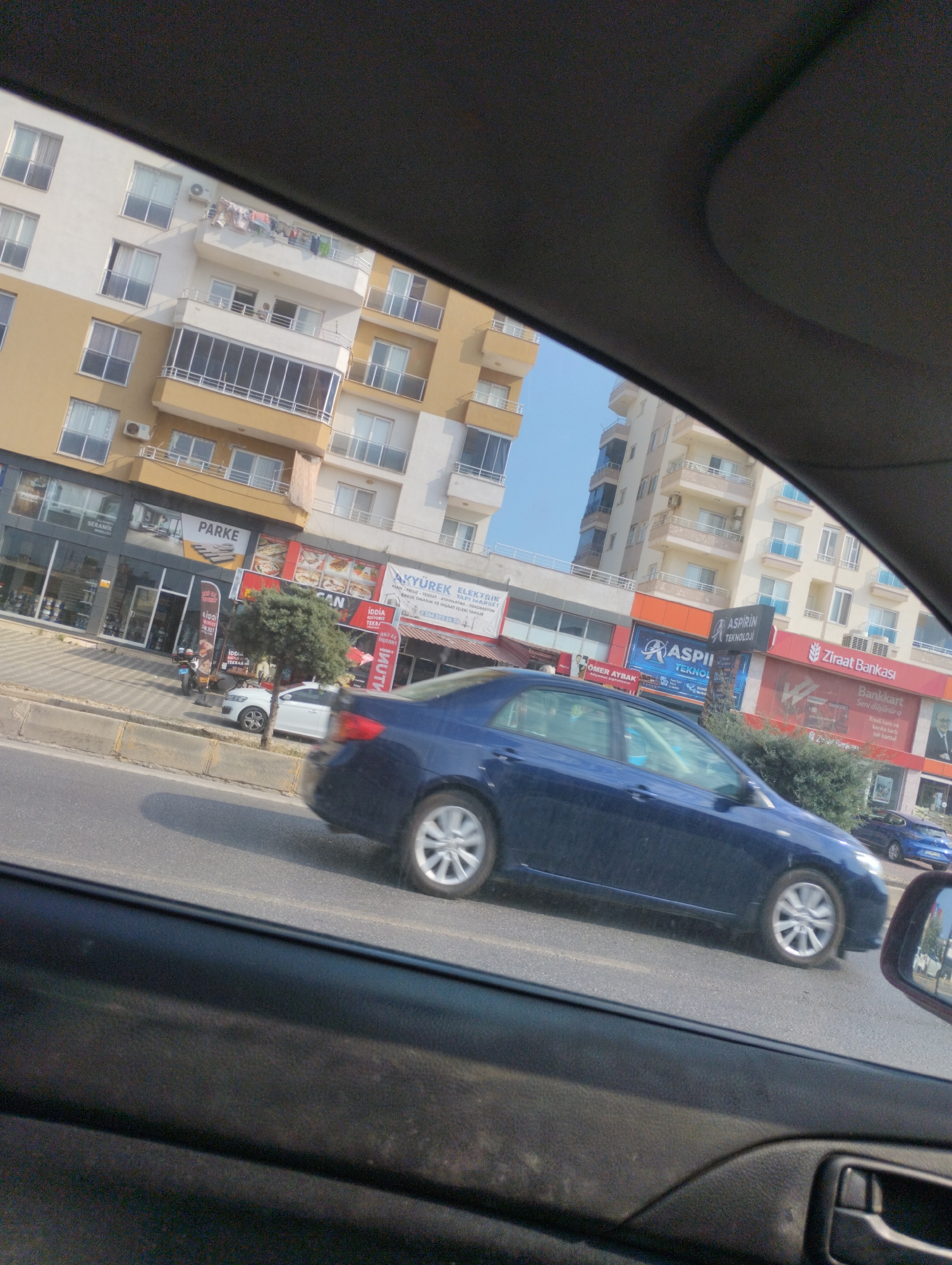 The image captures a street scene in Erdemli, Turkey, viewed from inside a vehicle. On the left, modern multi-story buildings with balconies and windows line the street. Several businesses are visible on the ground floor, including a store with a sign that reads "PARKE," indicating flooring or a similar product. Other shops with various signage are also present. A dark blue sedan is driving on the road in the foreground, with its wheels blurred due to motion. A white car is parked on the side of the road, and a small tree is situated between the road and the buildings. Further down the street, more buildings and a blue car are visible. The sky is clear and blue. The interior of the vehicle from which the photo is taken is partially visible, showing a dark dashboard and door panel.