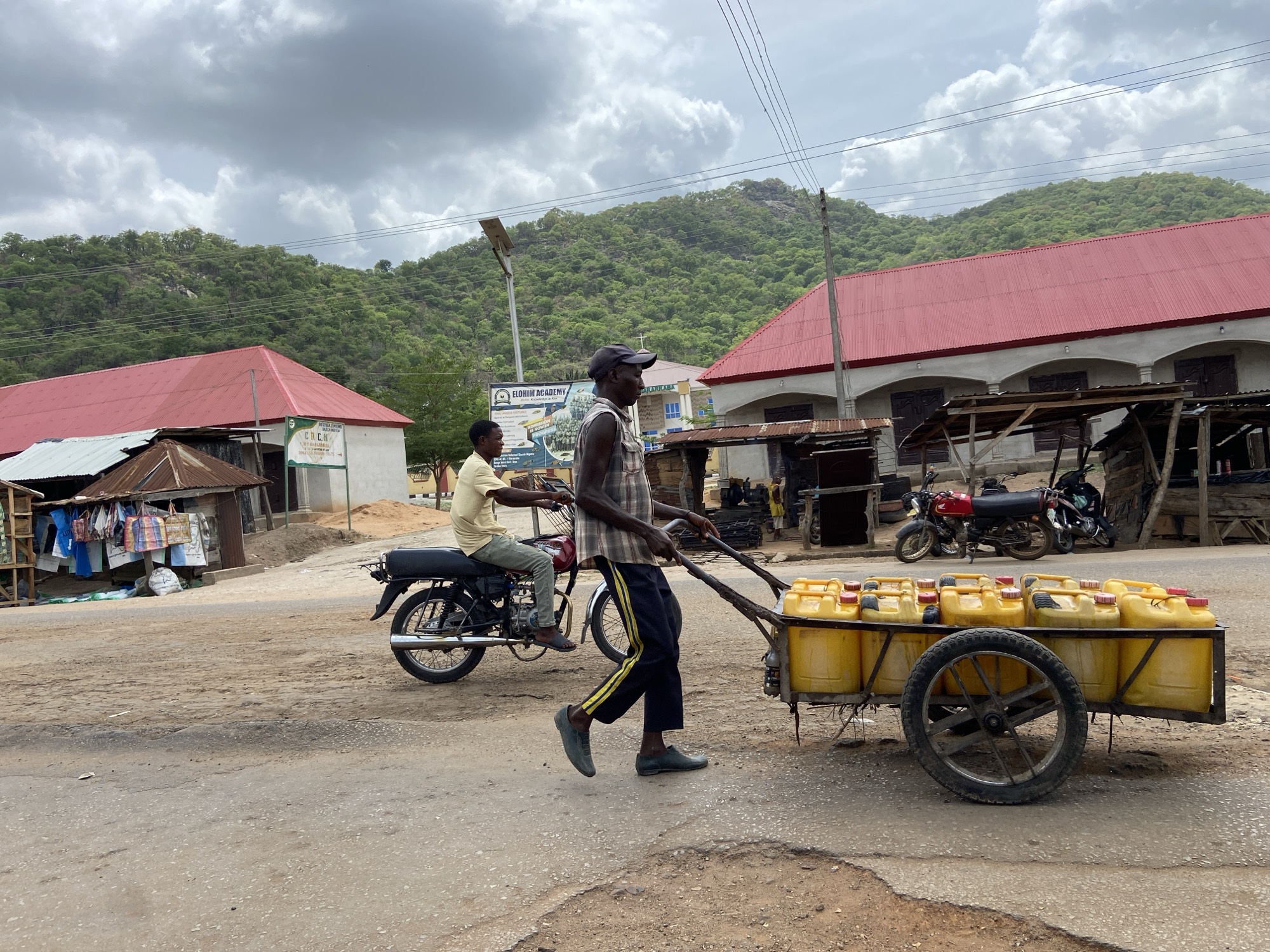 In Mararaba, Nigeria, a man in a plaid shirt and black cap is pulling a two-wheeled cart loaded with yellow jerrycans down a paved road. He wears dark pants with a yellow stripe and dark shoes. To his left, a man rides a black motorcycle. Several buildings with red roofs line the street, and a green, forested hill is in the background under a cloudy sky. Several motorcycles are parked in the background.