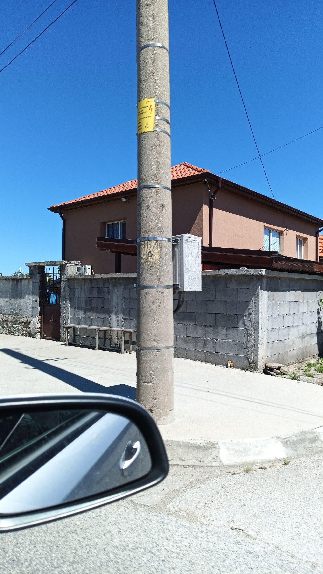 The scene depicts an outdoor view from a car in Boliartsi, Bulgaria. A concrete utility pole stands in the foreground, featuring a yellow warning sign and a grey metal box attached to it. Overhead power lines stretch across the clear blue sky. Behind the pole, a residential area is visible, consisting of a one-story brown house with a red tile roof. The house is partially obscured by a grey concrete block wall and a wrought iron gate. A simple wooden bench sits adjacent to the wall. The car's side mirror is visible in the lower-left corner of the frame, providing context for the viewpoint. The road and sidewalk are also visible, indicating an urban or suburban environment. No people are present in the frame.