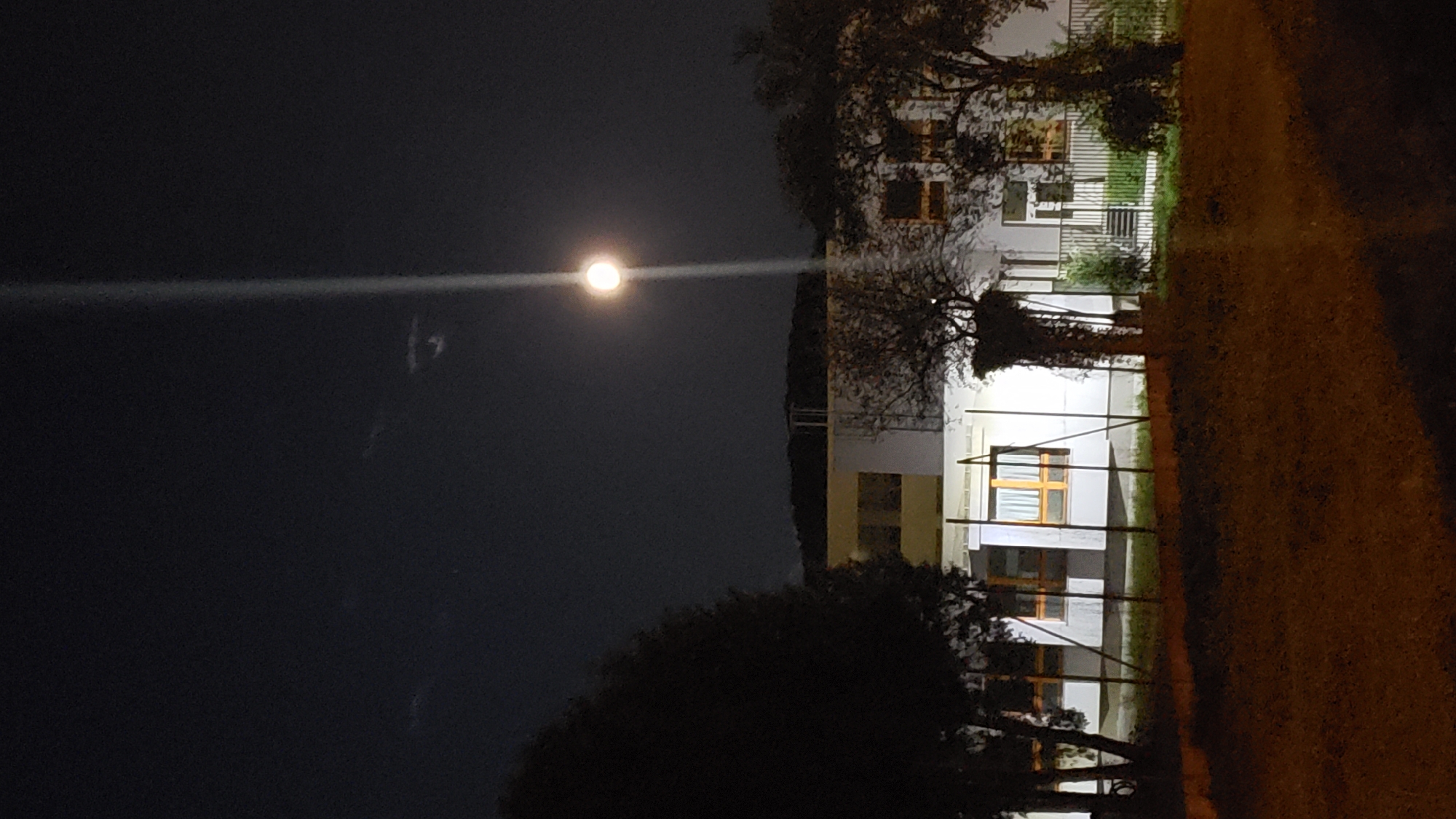 An exterior night-time vertical full shot captures a building, possibly a school or community center, illuminated by the moon and a bright overhead light. The building is a single-story structure with a light-colored facade and several windows. The moon is visible in the upper right quadrant of the sky, with a strong vertical light beam emanating from an unseen source, appearing to strike the building from above. Trees with dark foliage are present on either side of the building, and a fence runs along the foreground. The ground in front of the fence is covered in grass and appears somewhat unkempt. The sky is dark, with some faint cloud formations visible. The overall impression is a quiet, somewhat stark nighttime scene. There are no people or animals visible in the image. The location is Prešov, Slovakia, though the specific site within the city is not identifiable from the image alone.