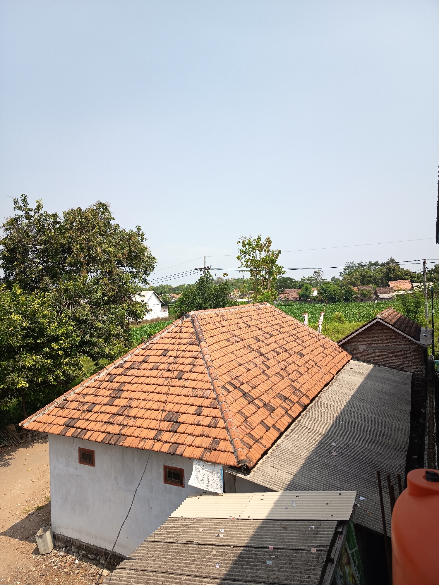 The image, taken from an elevated perspective, depicts a view of residential buildings and vegetation under a clear blue sky in Gebangan, Indonesia. In the foreground, a house with a tiled orange roof is visible, with white walls and two small, dark-framed windows. Adjacent to this house, another building with a corrugated metal roof casts a shadow. Further in the background, more houses with reddish-brown tiled roofs are scattered amongst lush green trees and fields of crops. A large, leafy tree dominates the left side of the frame. Power lines are strung across the scene, supported by utility poles. The overall impression is one of a rural or semi-rural Indonesian village setting on a bright, sunny day.