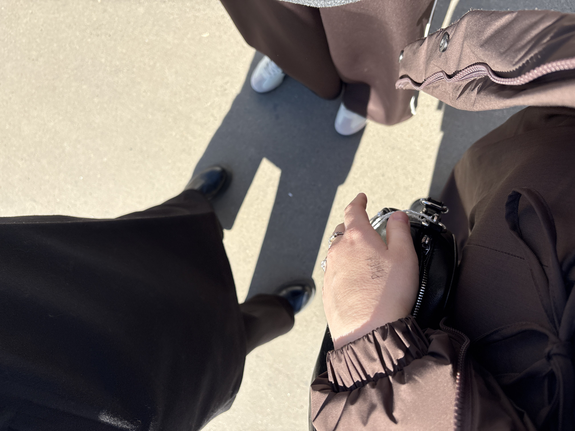 From an overhead viewpoint, the image depicts three people standing on a concrete surface, possibly a sidewalk or paved area, under daylight conditions. Two individuals are wearing long, dark-colored garments that extend to their shoes. One person is wearing white sneakers, while the other two have on black shoes. One person is wearing a brown jacket with a zipper. The photo appears to be taken in a city environment. While the picture doesn't definitively confirm it's Kazan, Russia, it aligns with urban environments that could exist there.