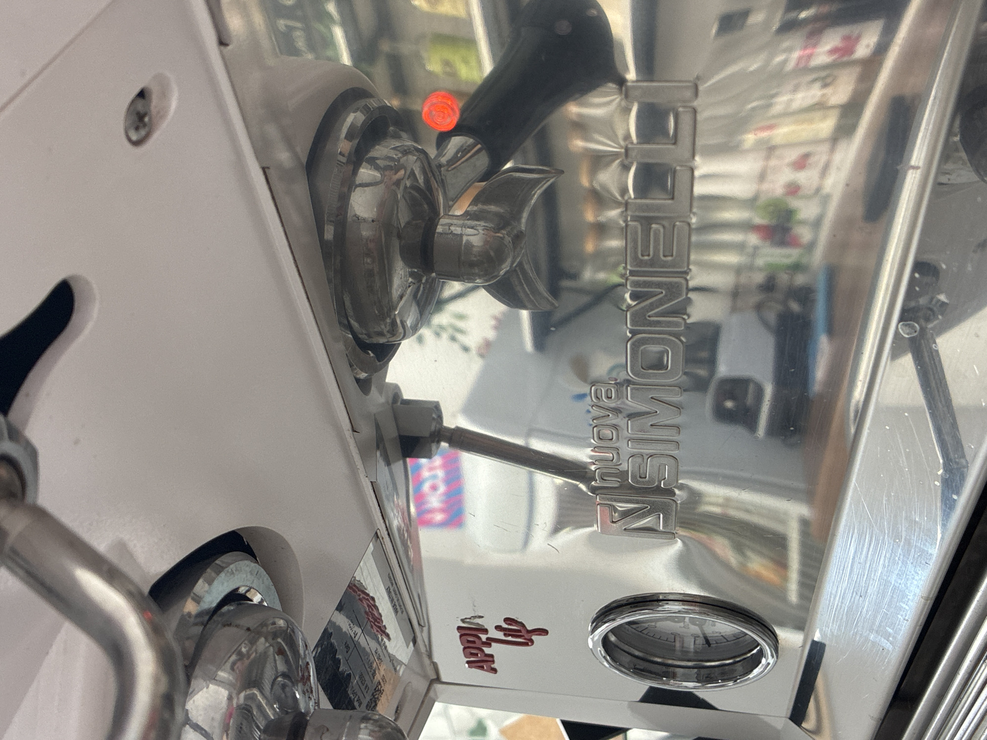 This image shows a close-up of a Nuova Simonelli espresso machine. The machine is mostly white and silver, with chrome accents. A portafilter is attached to the group head on the left side of the image. The brand name "Nuova Simonelli" is vertically displayed in chrome letters on the right. The red “Appia Life” logo is visible below the brand name. The surrounding environment and location are not discernible from this image.