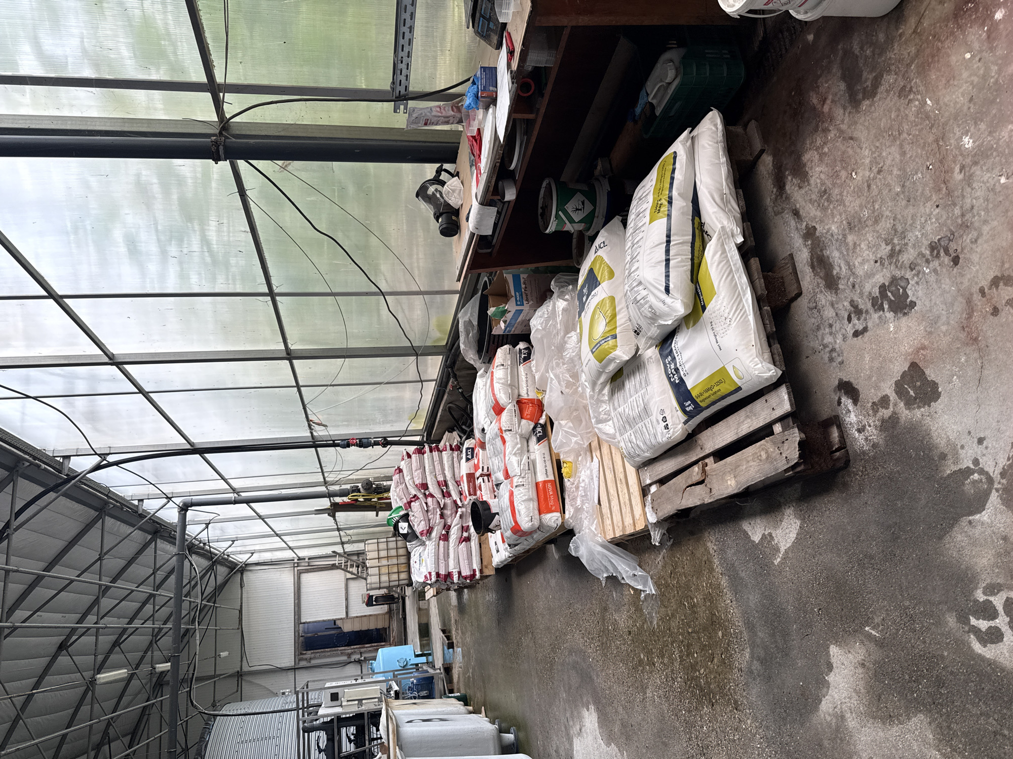 The image captures an indoor scene, likely within a greenhouse or a similar agricultural structure. The presence of metal framework and translucent roof panels suggest this. A variety of stacked fertilizer bags, some on wooden pallets, line the right side of the space, creating a visual barrier along the walkway. The bags are mostly white, with some having orange and red stripes or labels, and some that include the product names Nova Mag-N and Librel Fe-H17. A dark-colored table is present in the background, holding various supplies and equipment. The floor appears to be concrete with water stains and discoloration. In the distance, tanks and other equipment are visible, suggesting an area for water treatment or other processes. The image is taken indoors, there are no people shown, and there is no clear indication that this is located in Stara Zagora, Bulgaria.