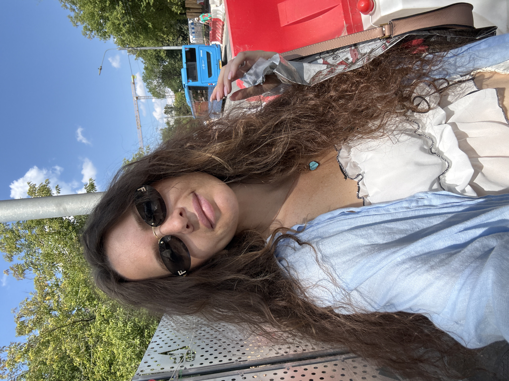 A young woman with long, wavy brown hair takes a selfie outdoors on a sunny day. She is wearing sunglasses and a light blue shirt over a white ruffled top. A light pink handbag is visible over her shoulder. The background shows a bright blue sky with some clouds, green trees, and construction equipment, including a red barrier and a blue truck. A crane is also visible in the distance. The woman appears relaxed and happy, with a slight smile. The image is oriented vertically.