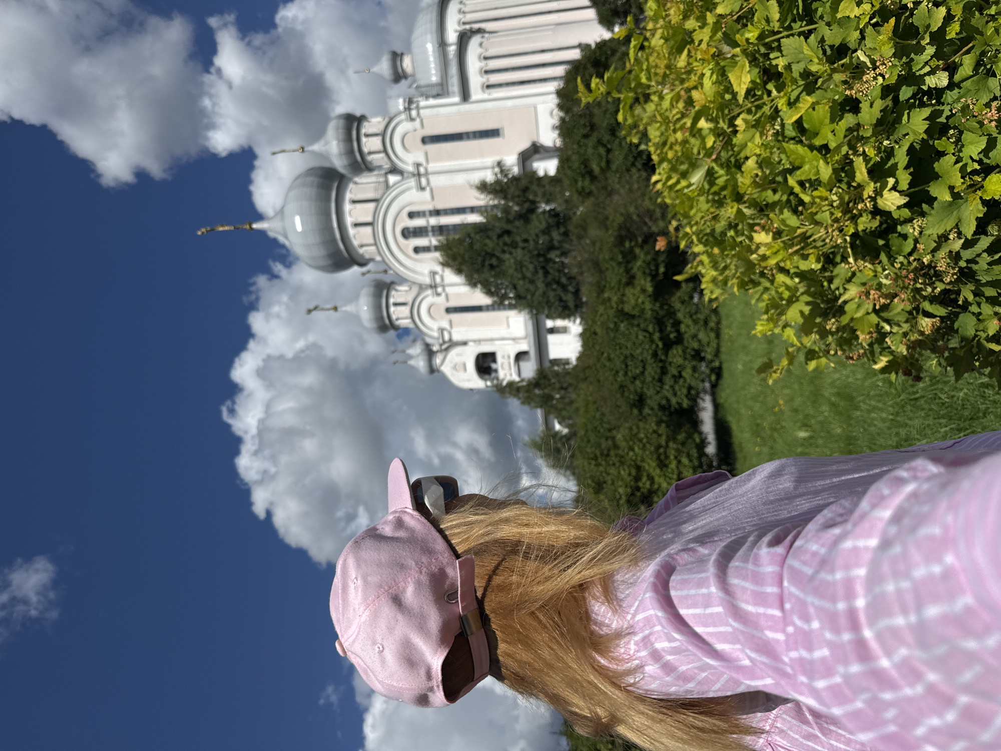 An outdoor, medium shot captures the back of a woman wearing a pink baseball cap and a pink and white striped shirt. Her blonde hair is visible beneath the cap, and she is wearing sunglasses. She is facing away from the camera, looking towards a large, white and light pink church with several silver domes. The church is situated behind a dense row of dark green trees, with bright green foliage in the foreground. The sky is a bright blue with fluffy white clouds. The overall scene suggests a sunny day. The architectural style of the church, with its multiple domes, indicates it is likely an Orthodox Christian church, possibly the Church of the Savior on Spilled Blood in Saint Petersburg, Russia, though confirmation would require further information.